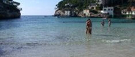 A beach with clear water, people in the water, and buildings on a hill in the background.