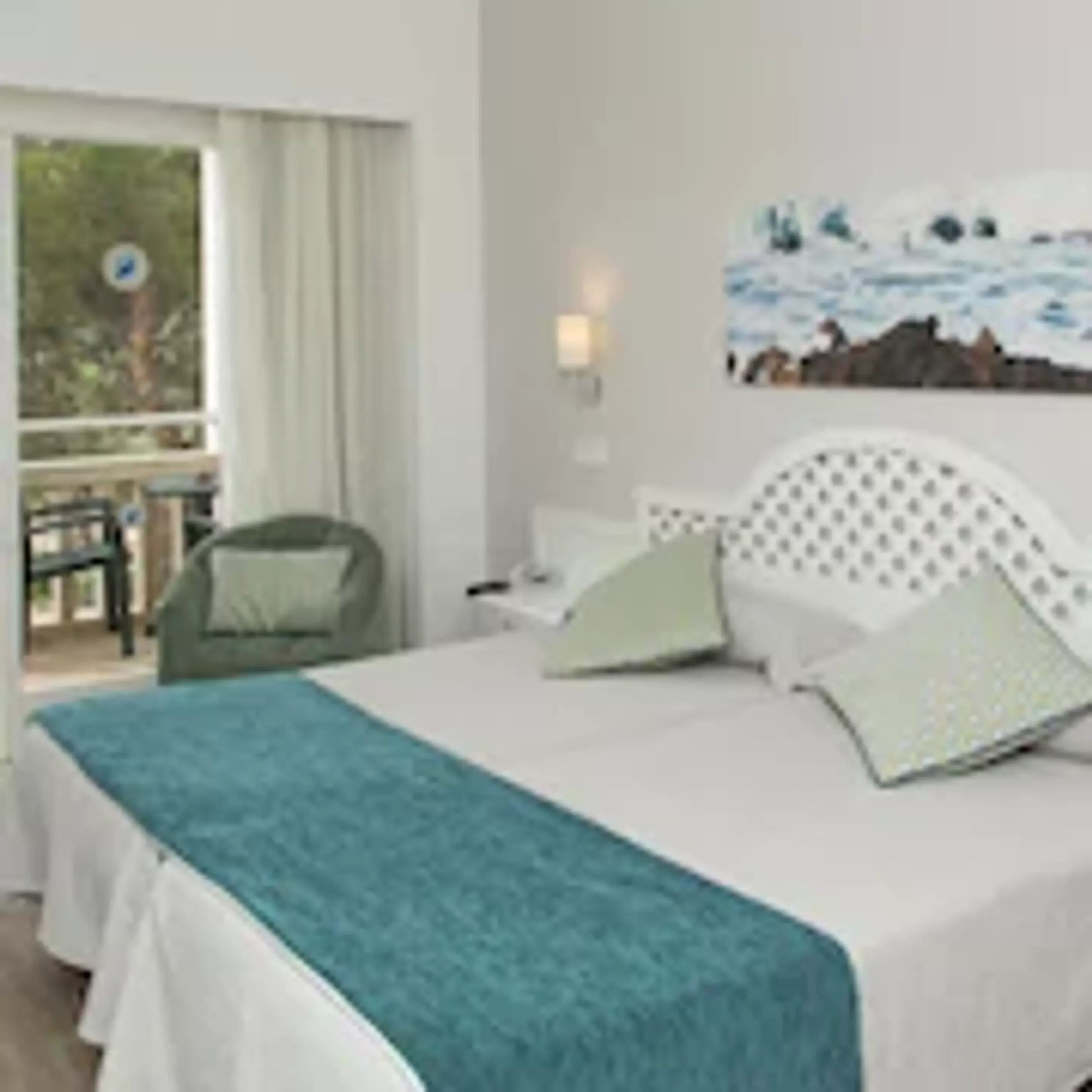 A hotel room with a bed, a window with curtains, a painting on the wall, and a chair outside the window.
