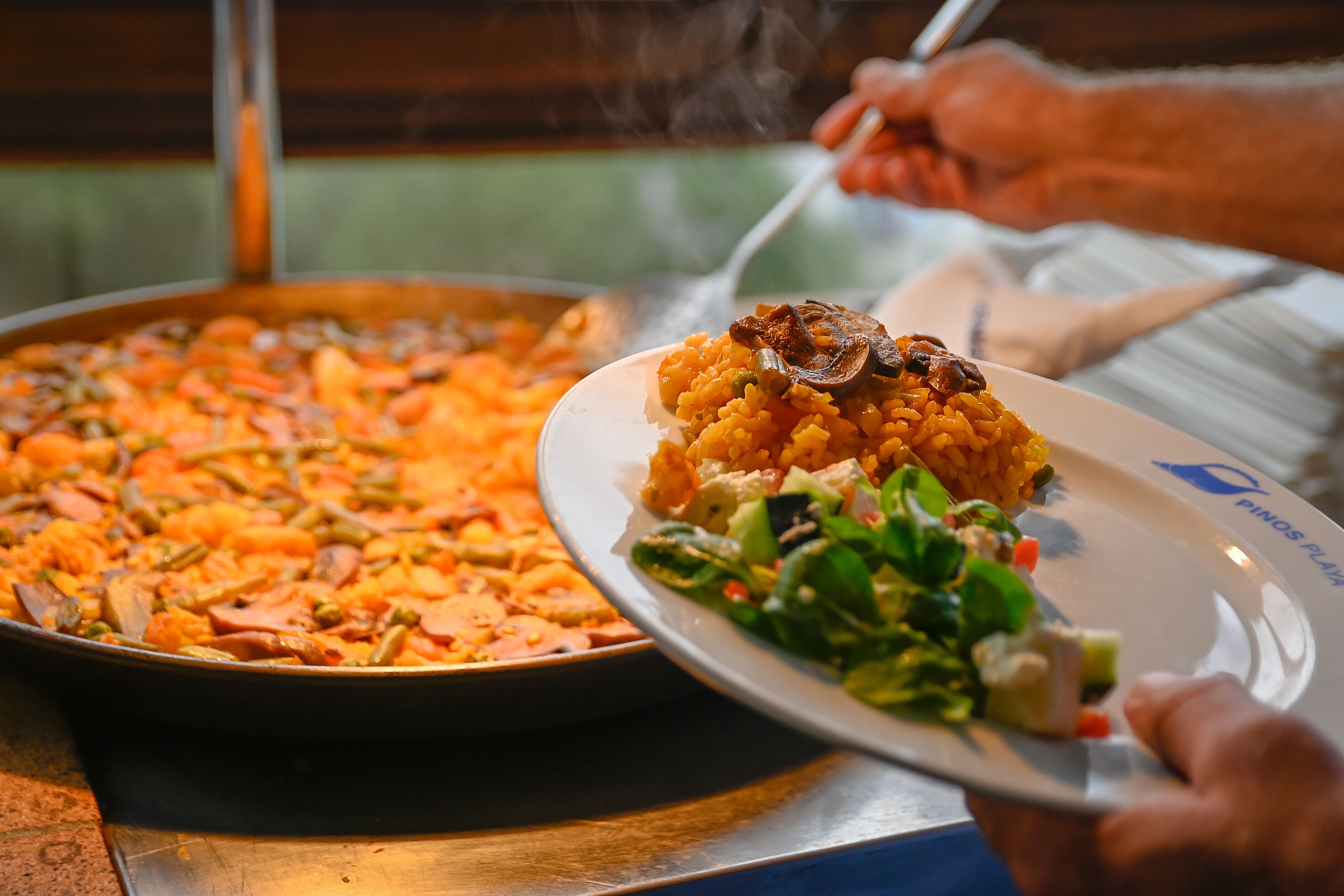 Une grande poêle remplie de nourriture dont la vapeur s’élève, une assiette avec une salade et la main d’une personne tenant une cuillère.