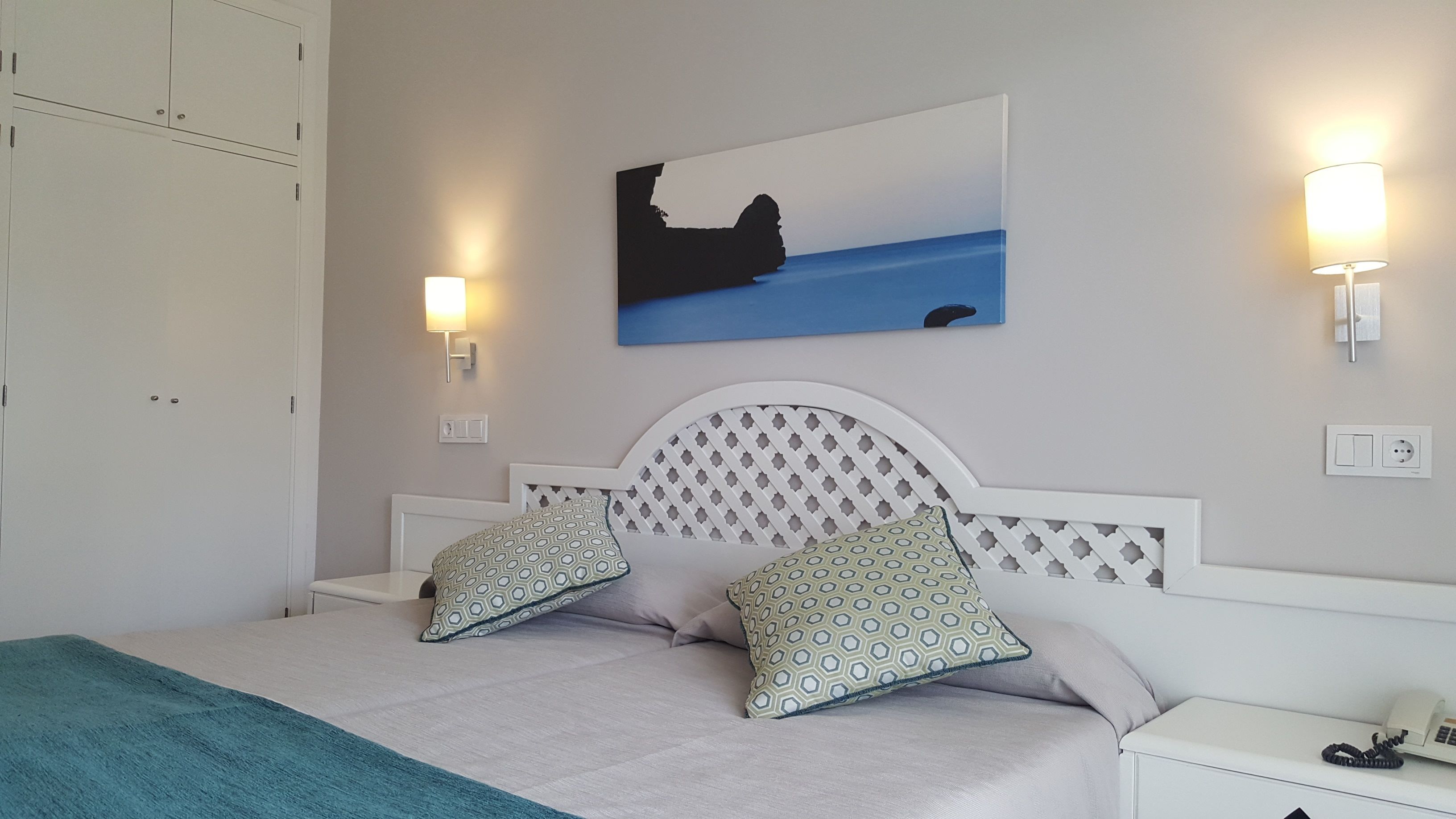 A bedroom with a bed, two patterned pillows, a headboard with a lattice design, a painting of a coastal scene, and bedside lamps.