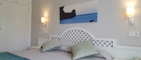 A bedroom with a bed, two patterned pillows, a headboard with a lattice design, a painting of a coastal scene, and bedside lamps.