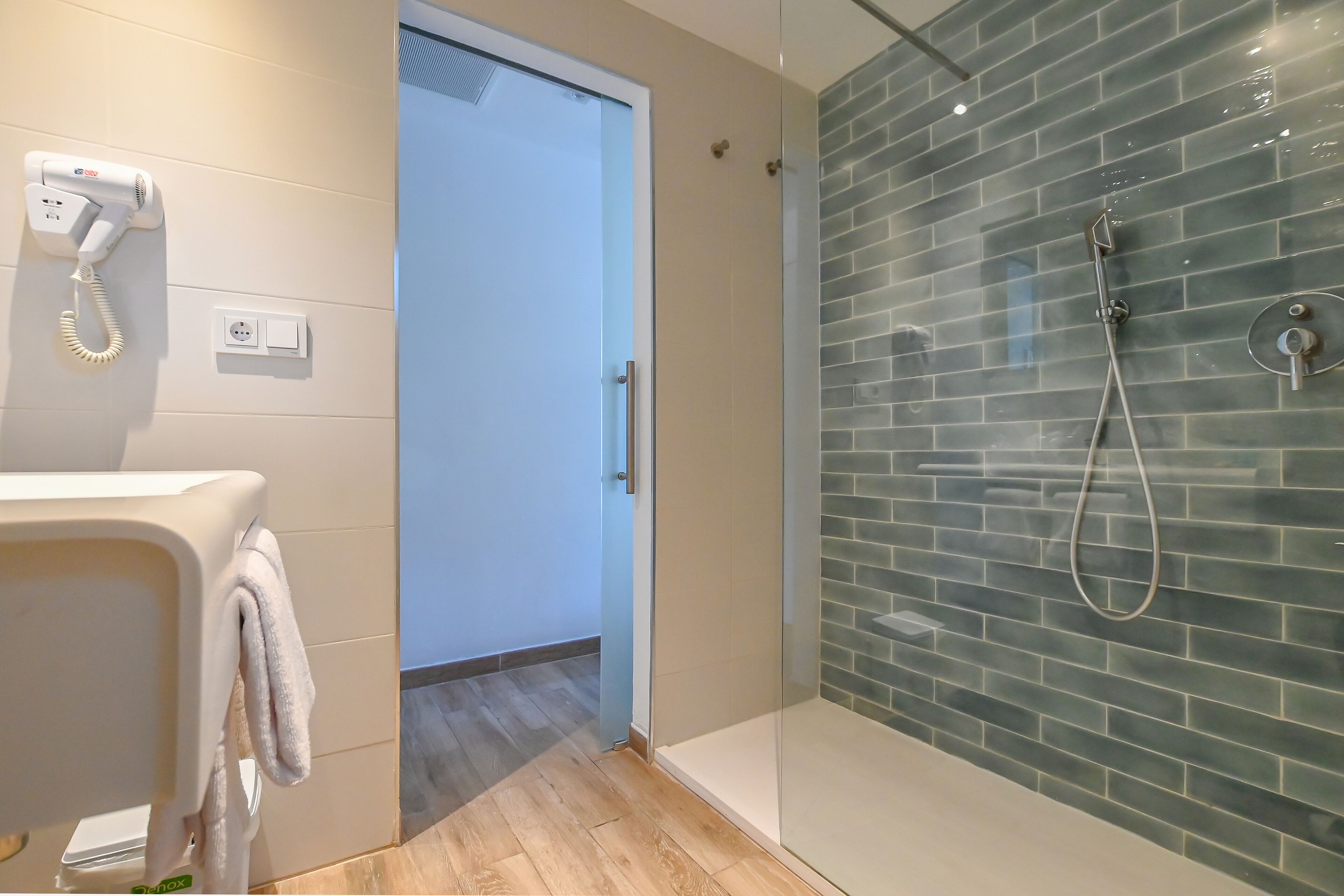 A modern bathroom with a glass shower enclosure, a hairdryer on the wall, and a towel hanging on the door.