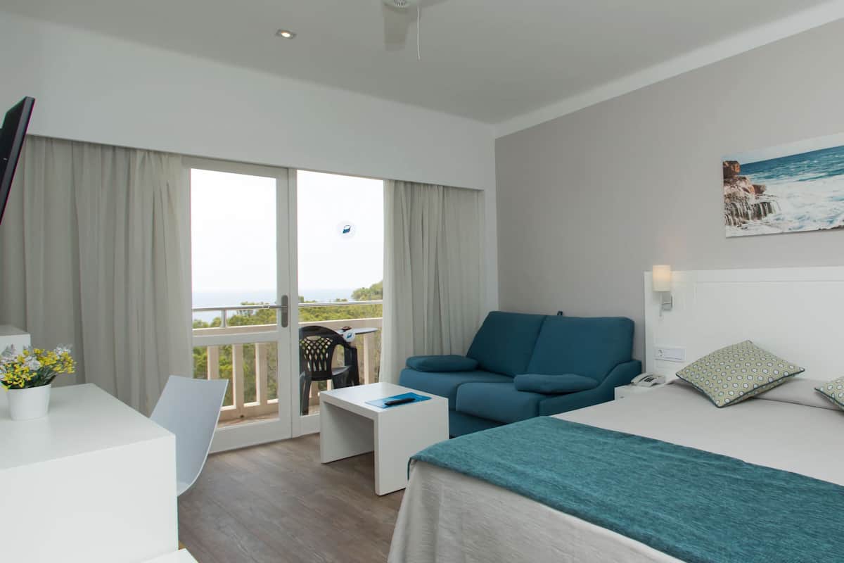 A modern hotel room with a balcony, a bed with a teal bedspread, a blue sofa, and a painting of a coastal scene.