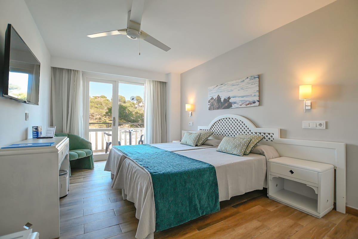 A hotel room with a bed, bedside tables, a desk, a chair, and a balcony with a view of trees.