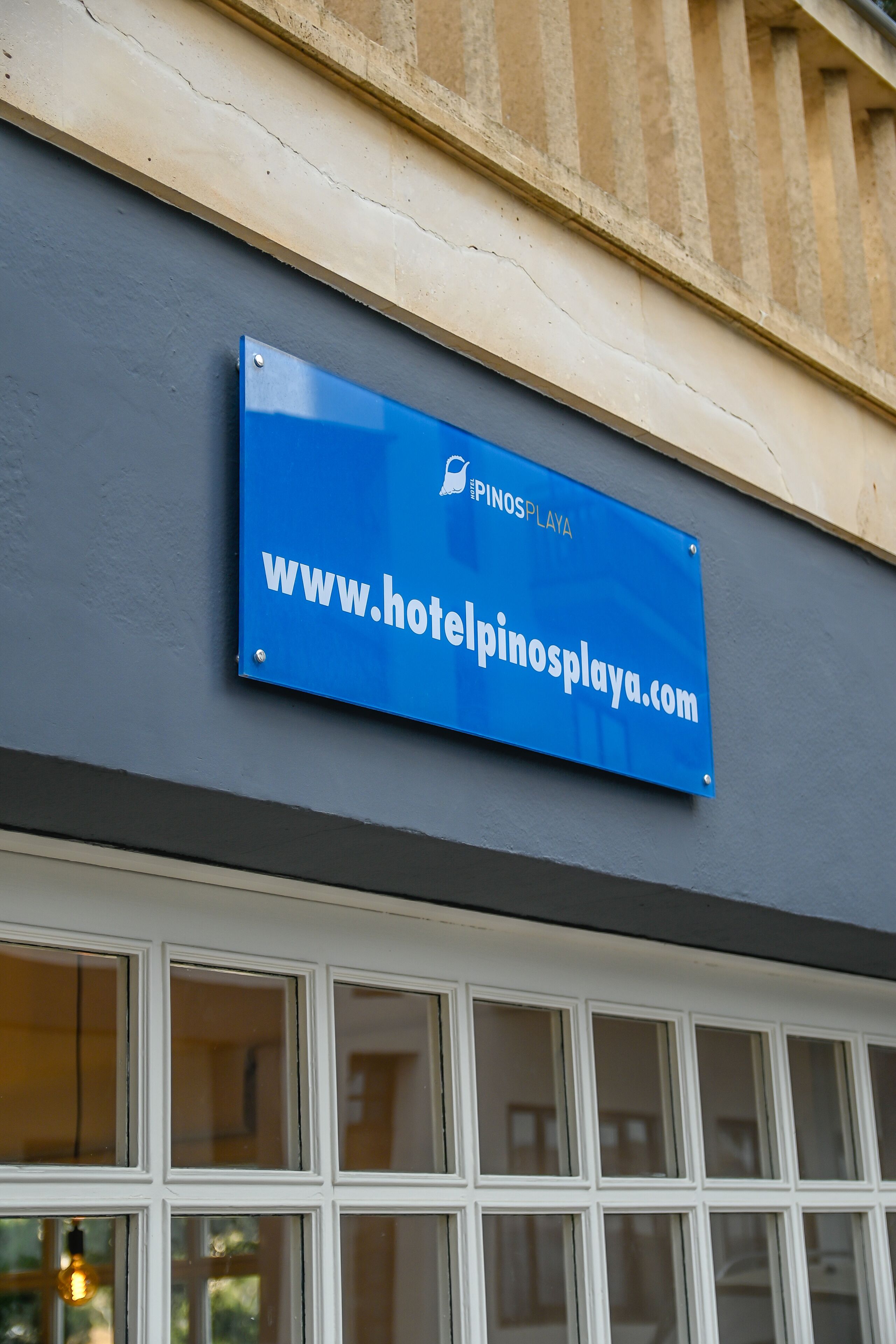 Un letrero azul con el texto "PINOS PLAYA" y "www.hotelpinosplaya.com" en la fachada de un edificio.