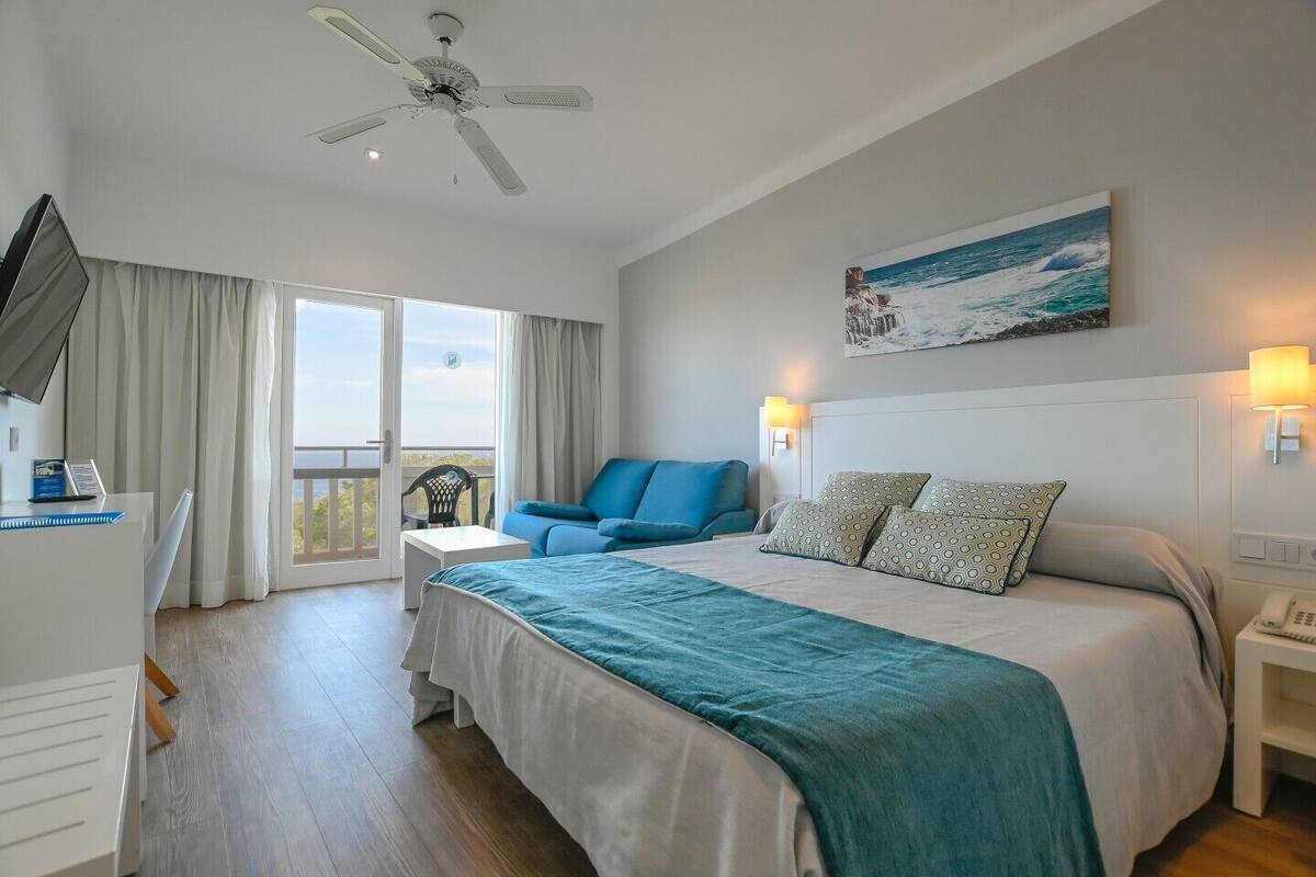 A bedroom with a large bed, a blue sofa, a ceiling fan, and a painting of a beach scene.