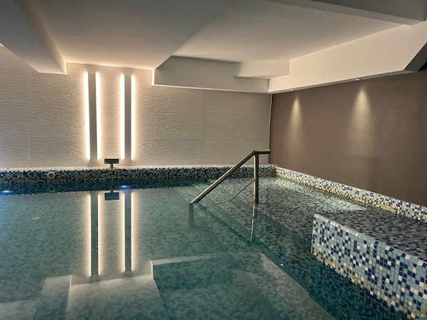 An indoor swimming pool with a tiled floor and walls, a metal handrail, and vertical light fixtures.