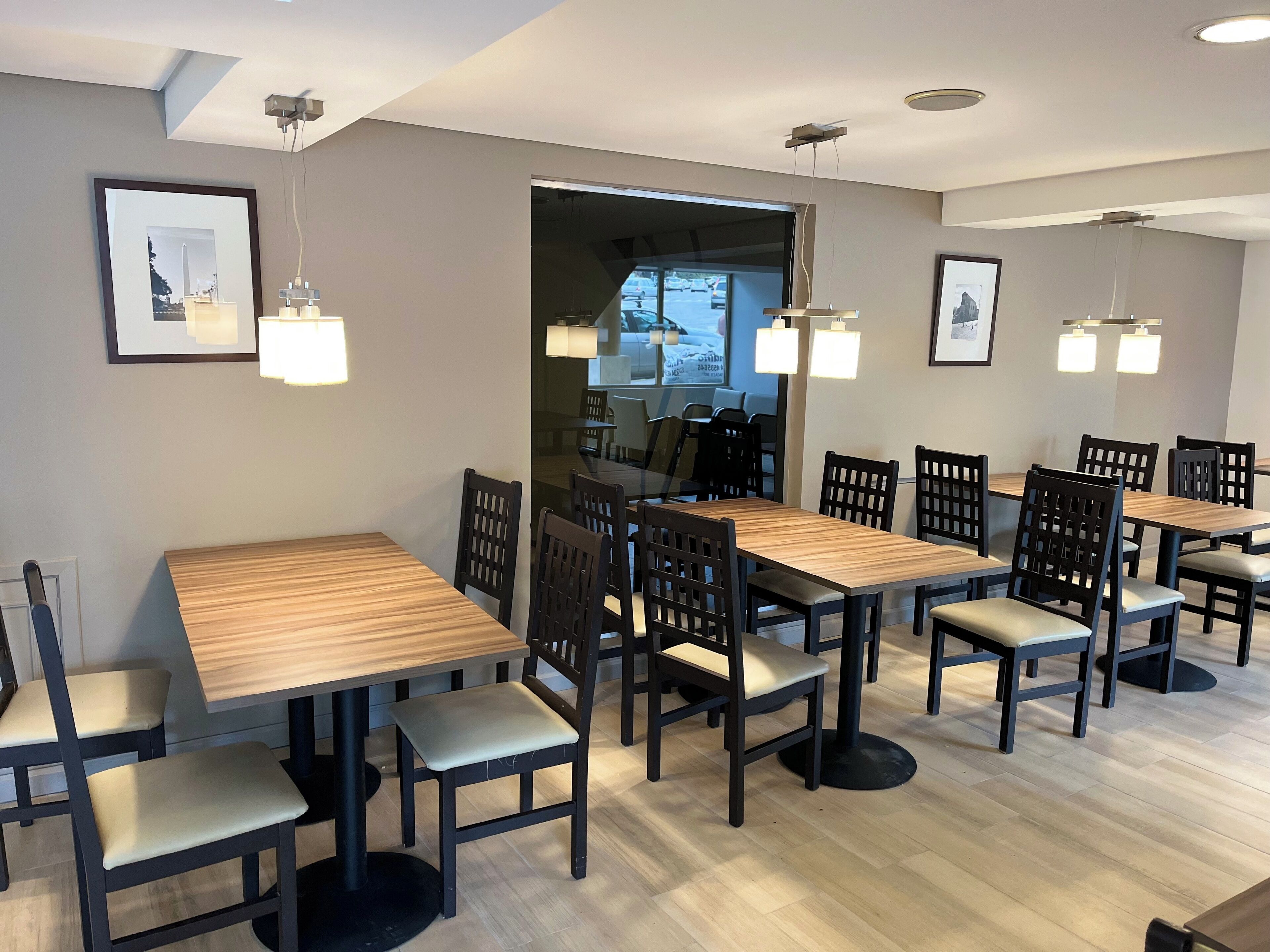 Un comedor con mesas de madera y sillas negras, cuadros en las paredes y luces empotradas en el techo.