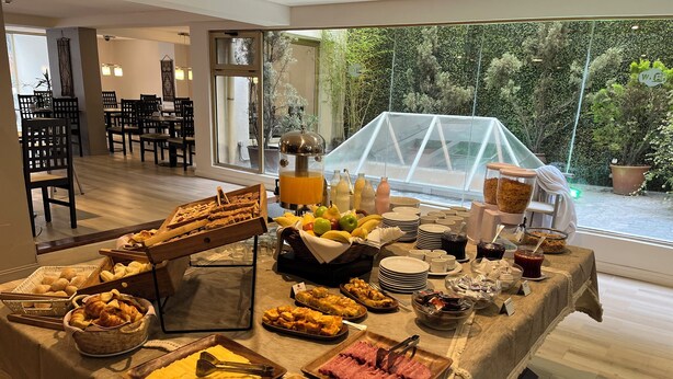 A buffet table with various food items, including pastries, fruits, and meats, set up in a room with large windows overlooking a garden. - Kenton Palace Bariloche (San Carlos de Bariloche)