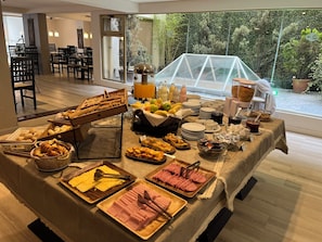 A buffet table with various food items, including pastries, fruits, and meats, set up in a room with large windows overlooking a garden. - Kenton Palace Bariloche (San Carlos de Bariloche)