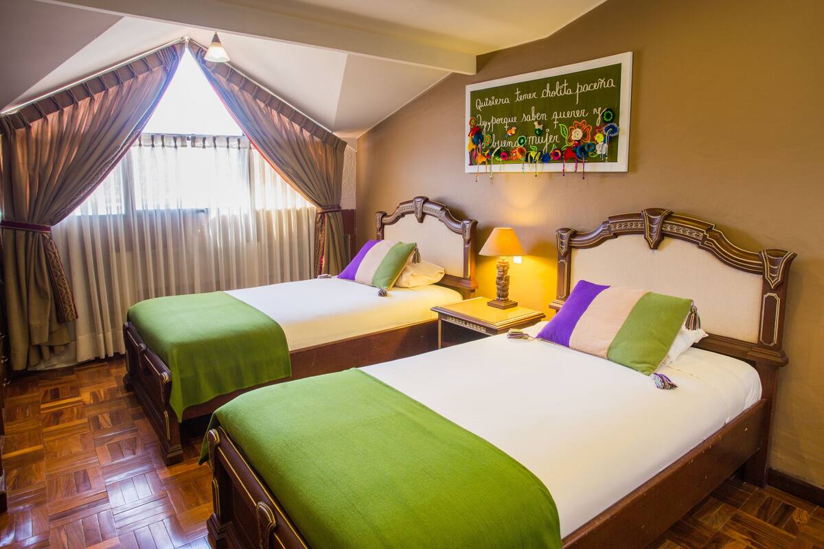 A hotel room with two beds, wooden flooring, a window with curtains, a bedside table with a lamp, and a wall decoration.