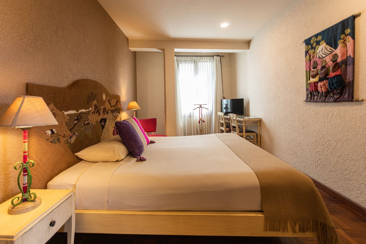 A hotel room with a bed, a nightstand with a lamp, a television, and a painting on the wall.