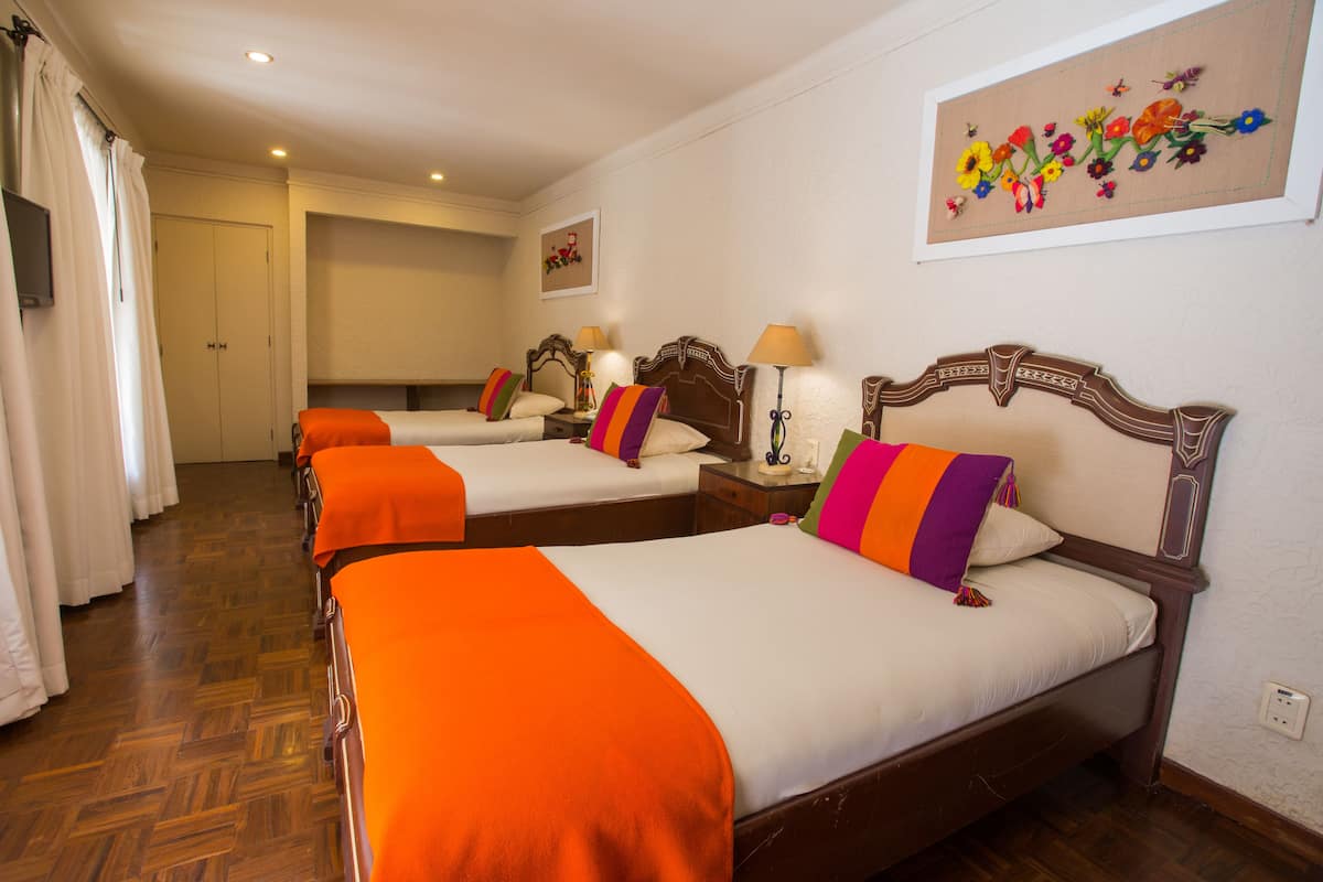 A hotel room with four single beds, each with an orange blanket and a wooden headboard. There is a painting of flowers on the wall, a wooden floor, and a door in the background.