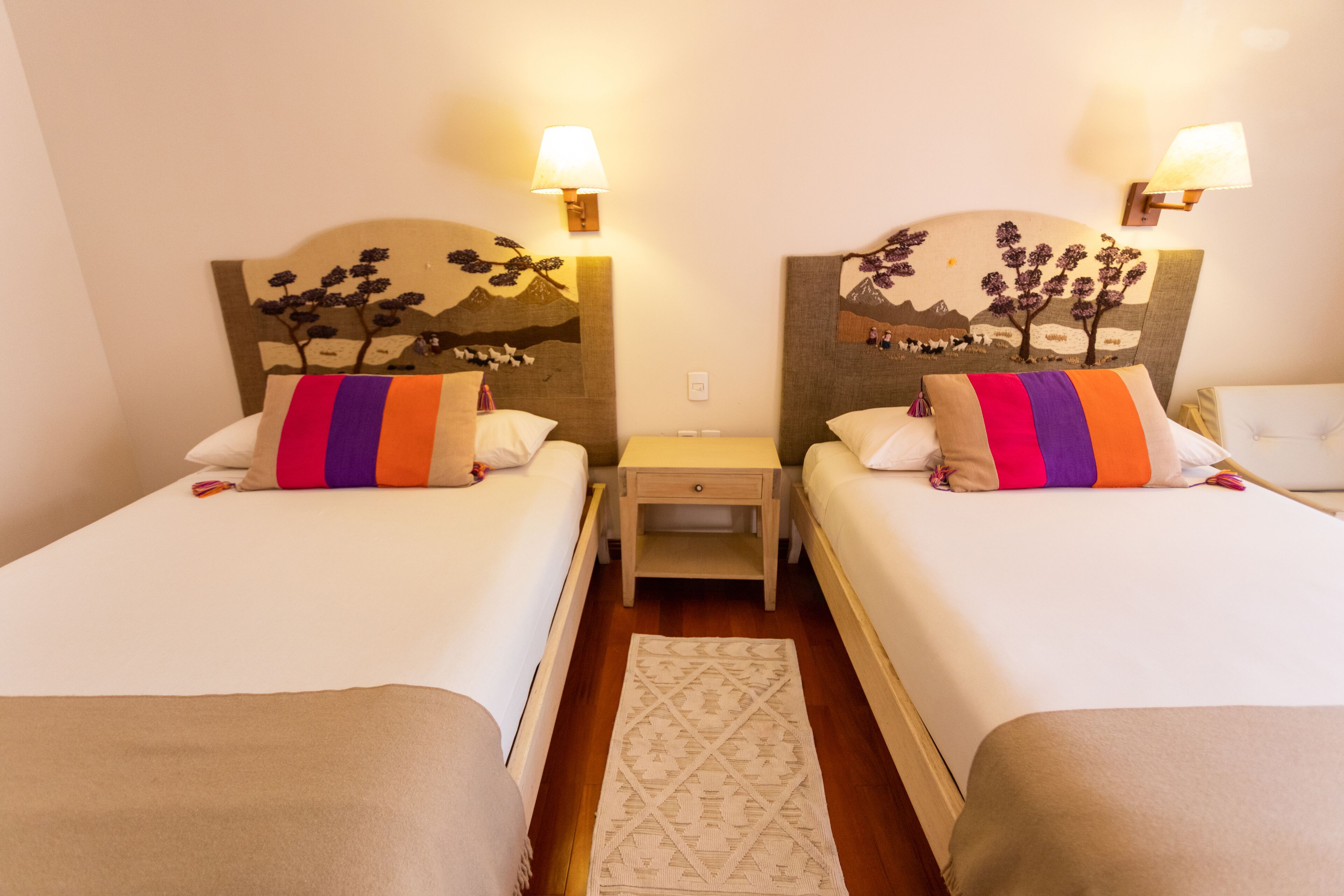 Una habitación de hotel con dos camas, cada una con un cabecero decorado con un diseño de árbol, una mesita de noche entre ellas y una pequeña mesa.