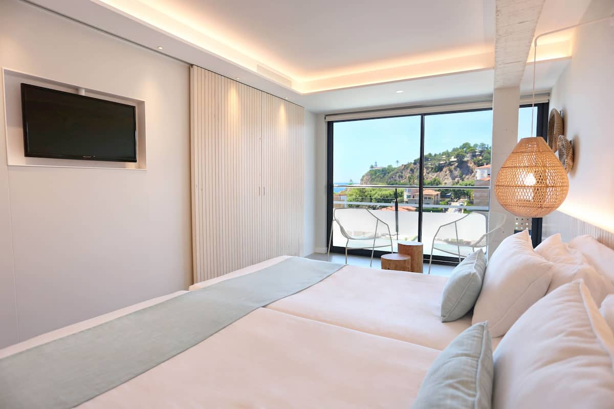 Une chambre moderne avec un grand lit, un téléviseur fixé au mur et une vue sur l’extérieur grâce à de grandes fenêtres.