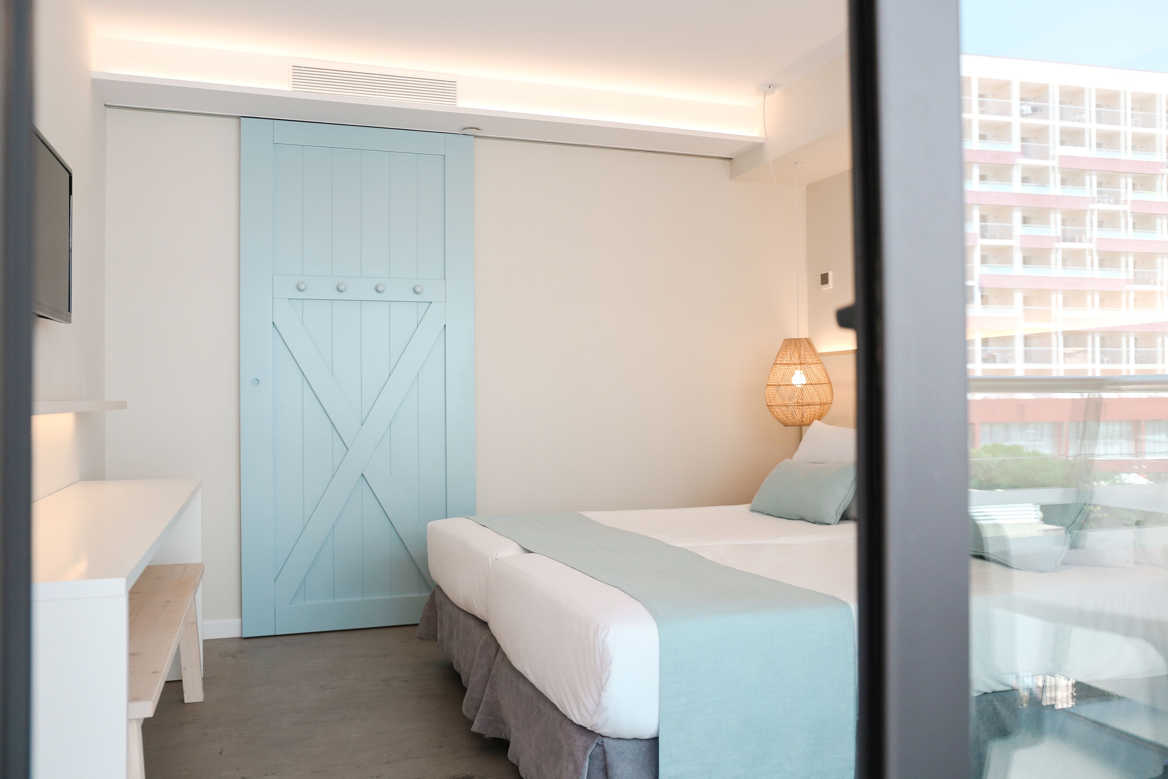 Un dormitorio con cama, escritorio, silla, armario y ventana con vista a edificios.