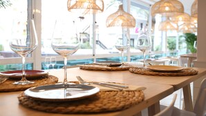 Una mesa de comedor dispuesta con platos, cubiertos y copas de vino, con individuales tejidos y lámparas colgantes.