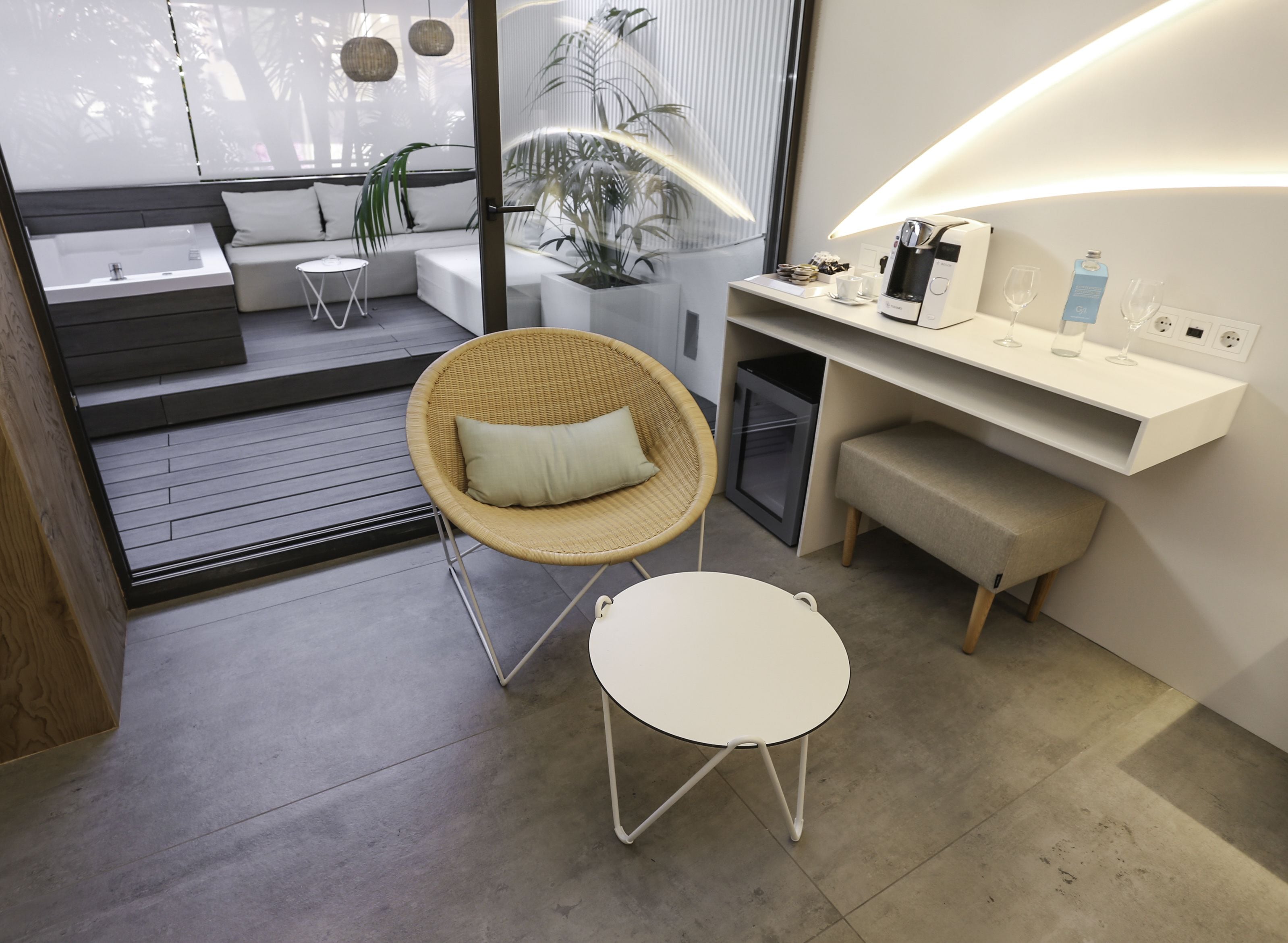 Et moderne, lyst rum med en siddeplads, bestående af en fletstol og et lille bord, en indbygget bænk, et hvidt konsolbord med kaffemaskine, samt et stort vindue med udsigt over grønne omgivelser.