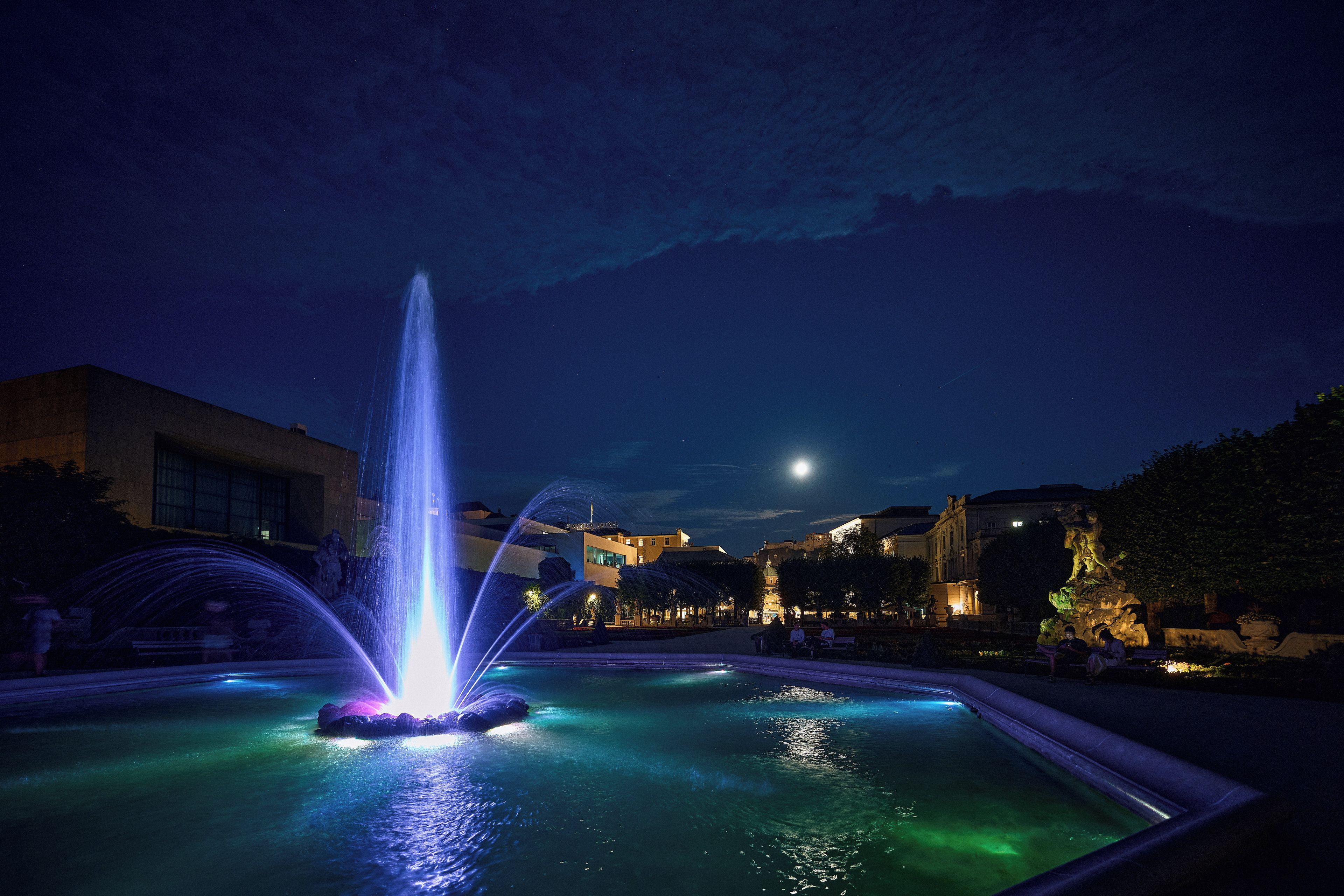 Ein nächtlicher Brunnen mit Wasserstrahlen, umgeben von einem Teich, mit Gebäuden und einem Mond im Hintergrund.
