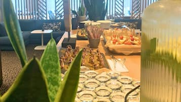 Un comedor con una mesa preparada para una comida, una planta grande en primer plano y vistas al exterior a través de una ventana con persianas verticales.