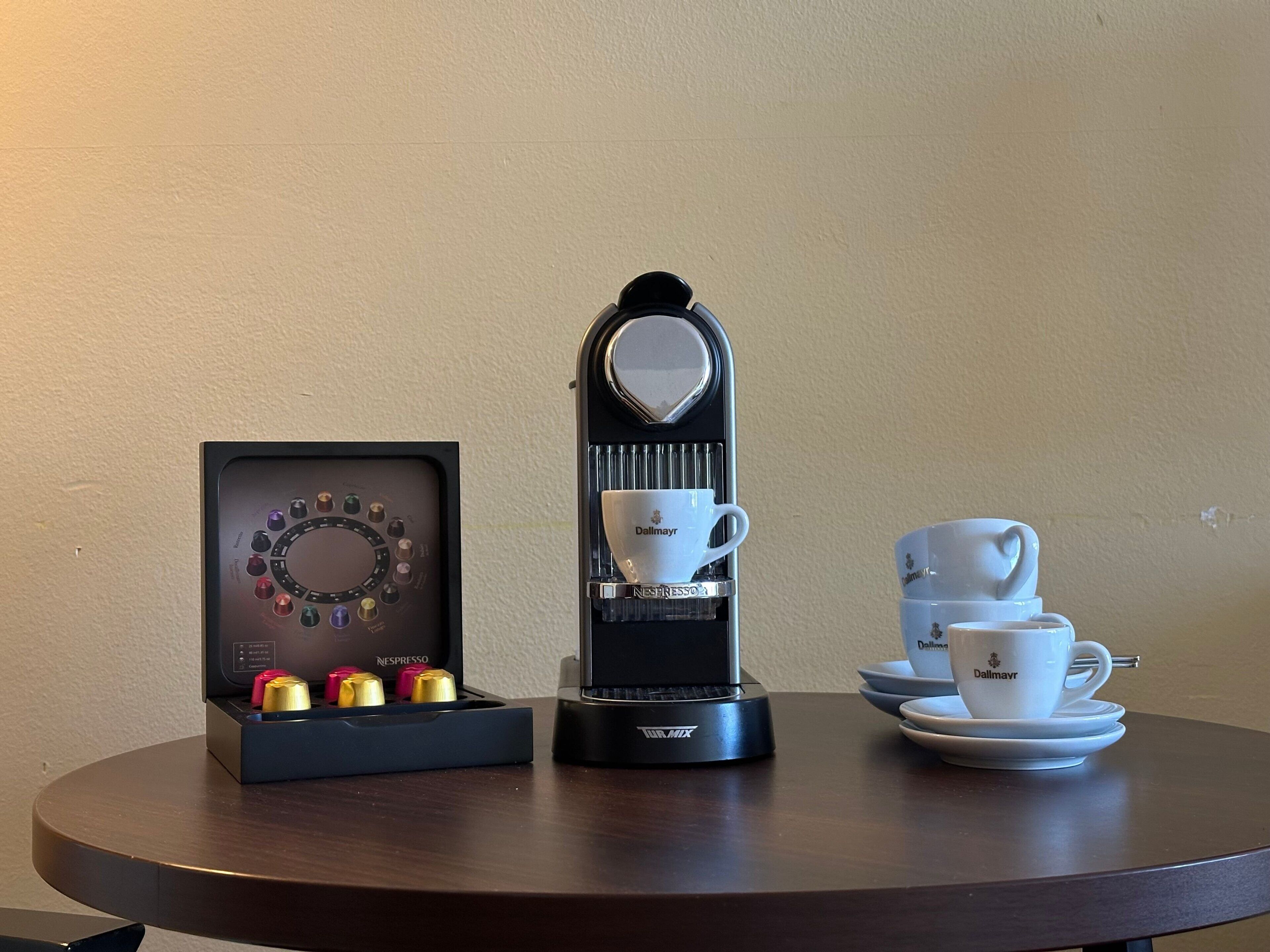 A coffee machine with a cup holder, a display case with coffee pods, and a set of white cups and saucers on a table.