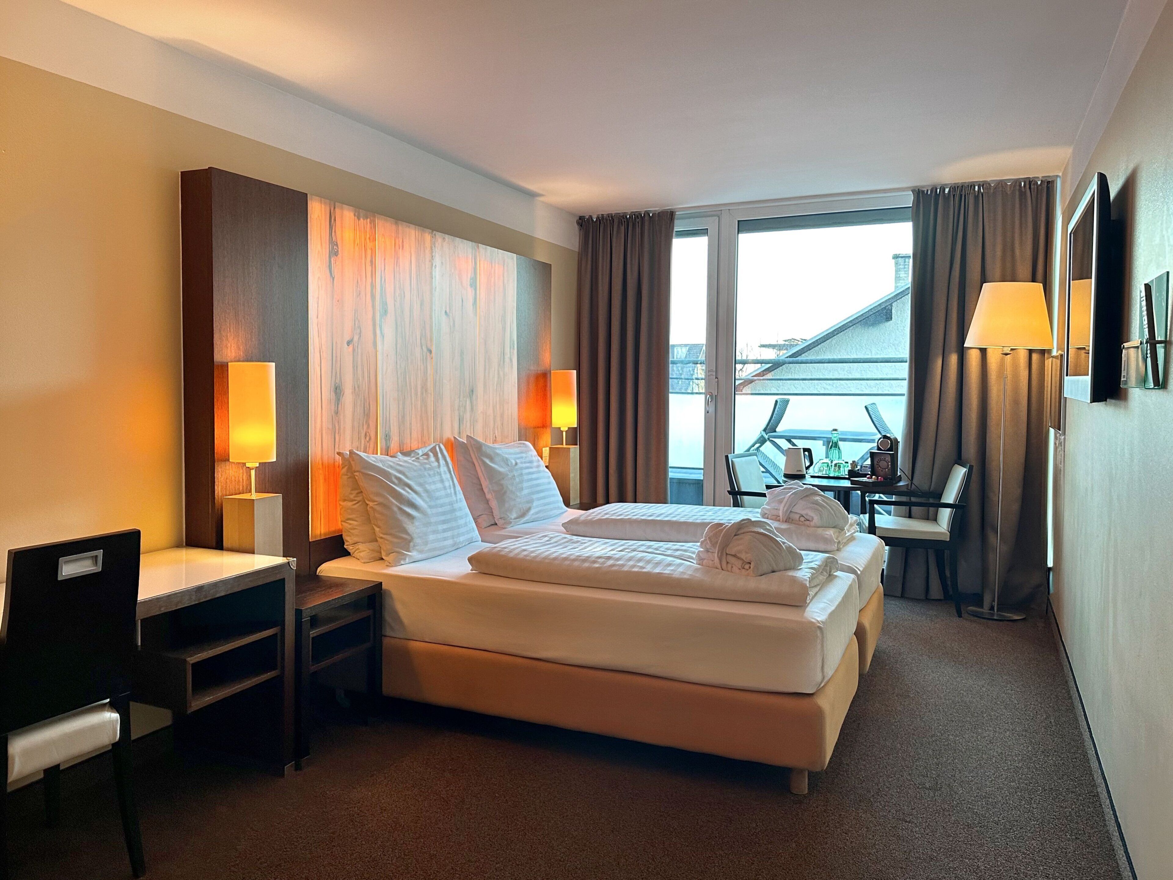 A hotel room with a large bed, two bedside tables with lamps, a desk with a chair, and a view of the outside through a large window.