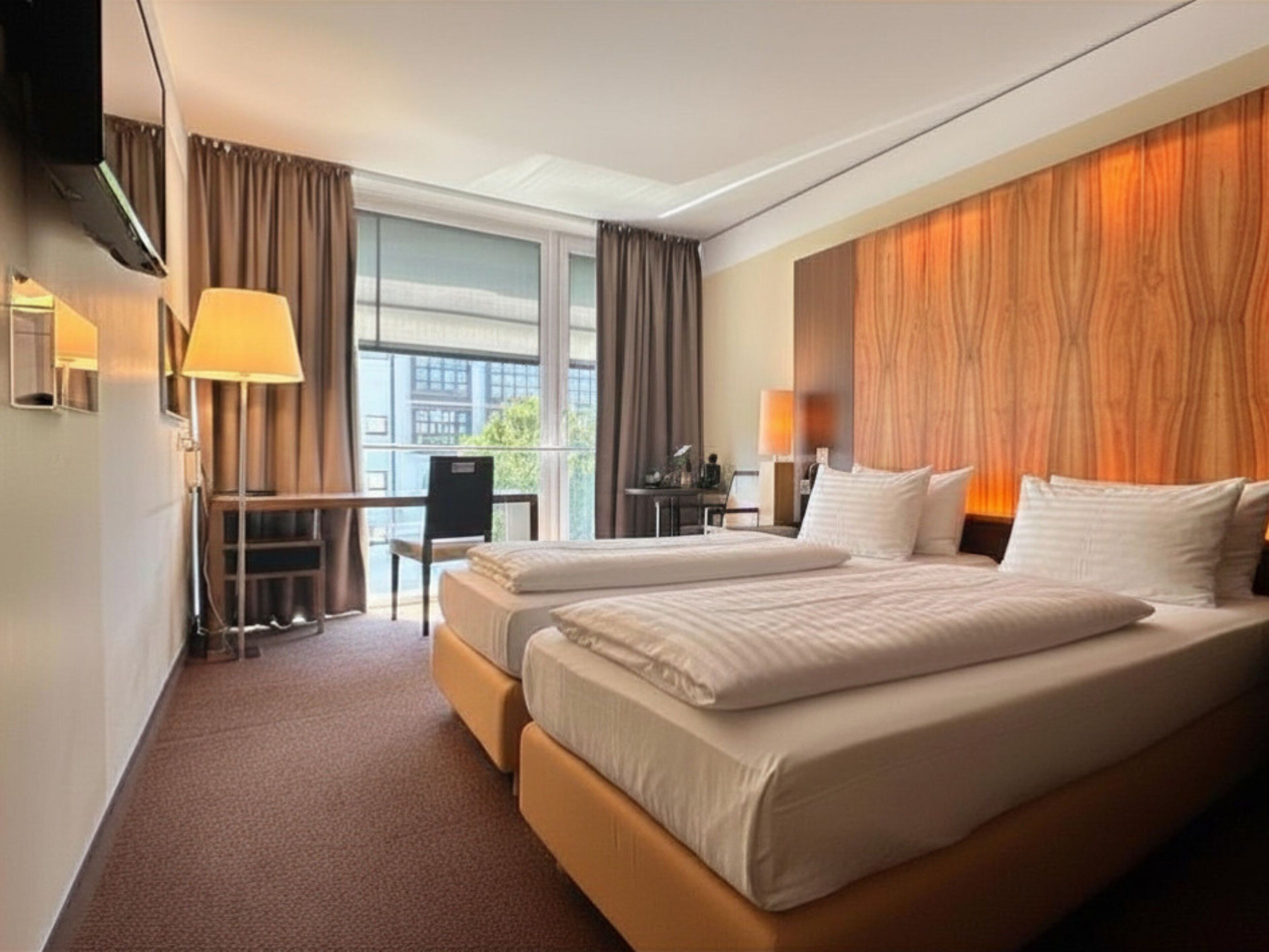 Quarto de hotel com duas camas, uma escrivaninha, uma cadeira e uma janela grande.