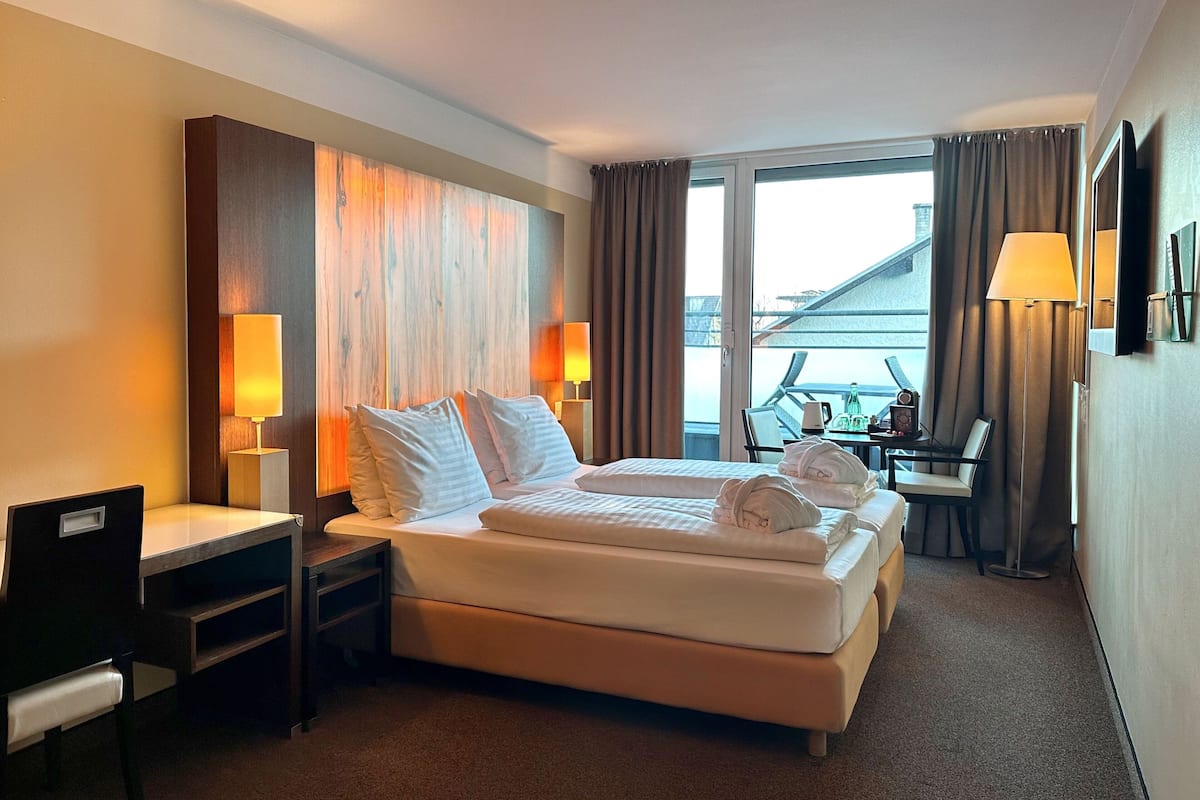 A hotel room with a large bed, two bedside tables with lamps, a desk with a chair, and a view of the outside through a large window.