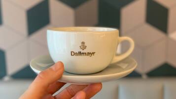 A hand holding a white cup with 'Dallmayr' text and logo, set against a geometric patterned background.