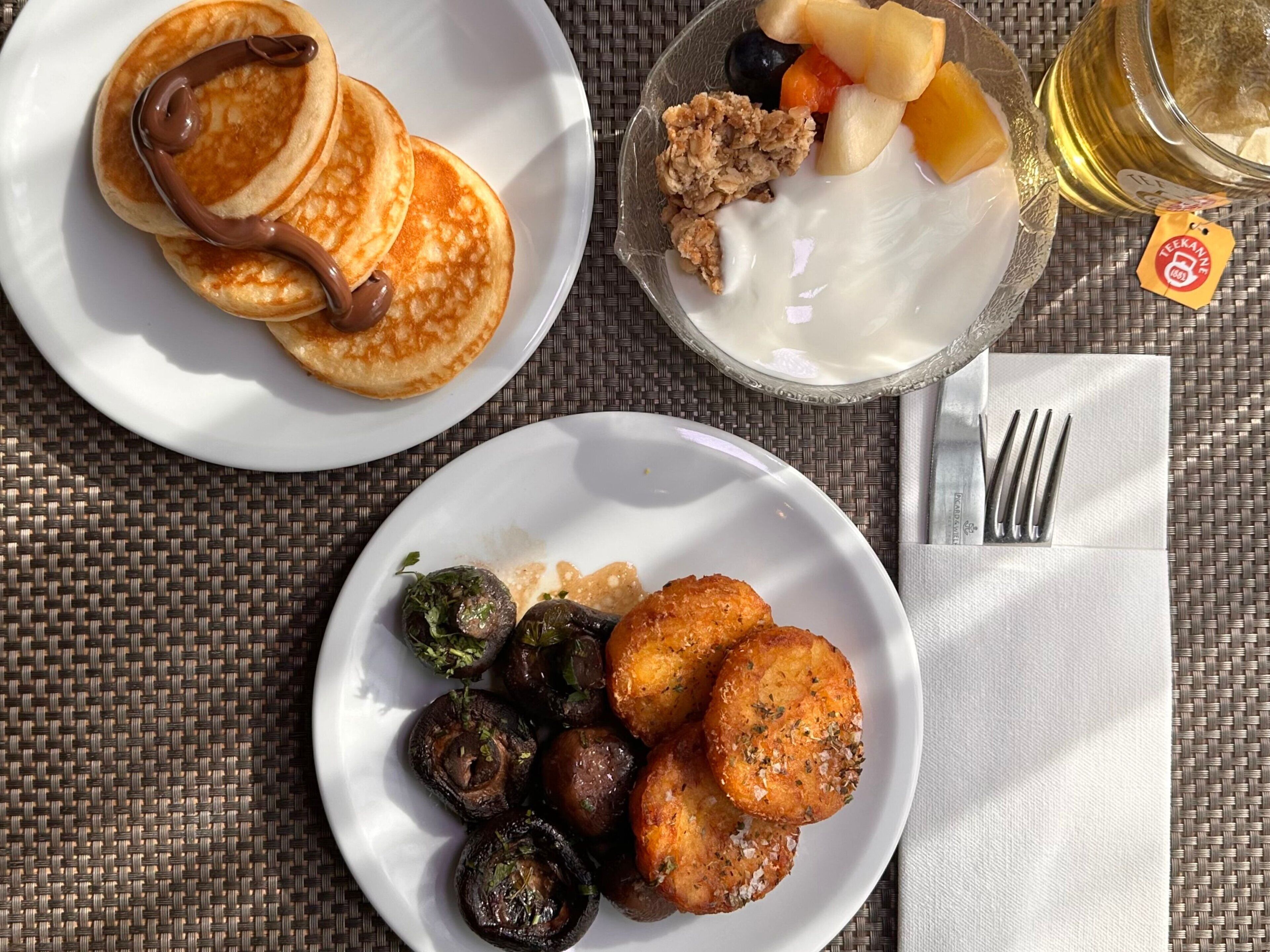 A breakfast setting with pancakes, fruit, yogurt, and roasted vegetables on a woven table.