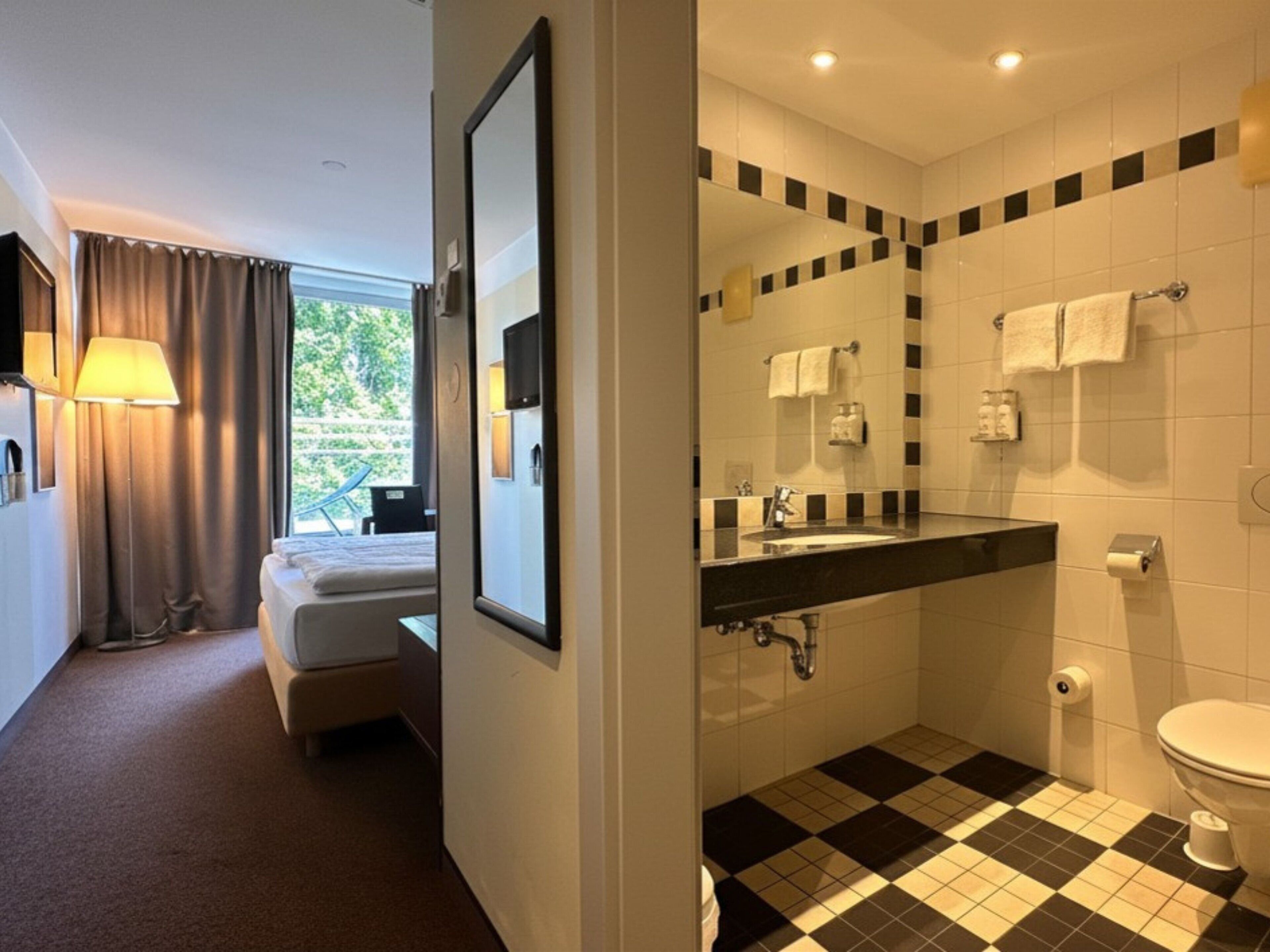 Hotelkamer met een bed, een bureau, een lamp, een raam met gordijnen en een badkamer met een zwart-wit betegelde vloer, een wastafel en een toilet.