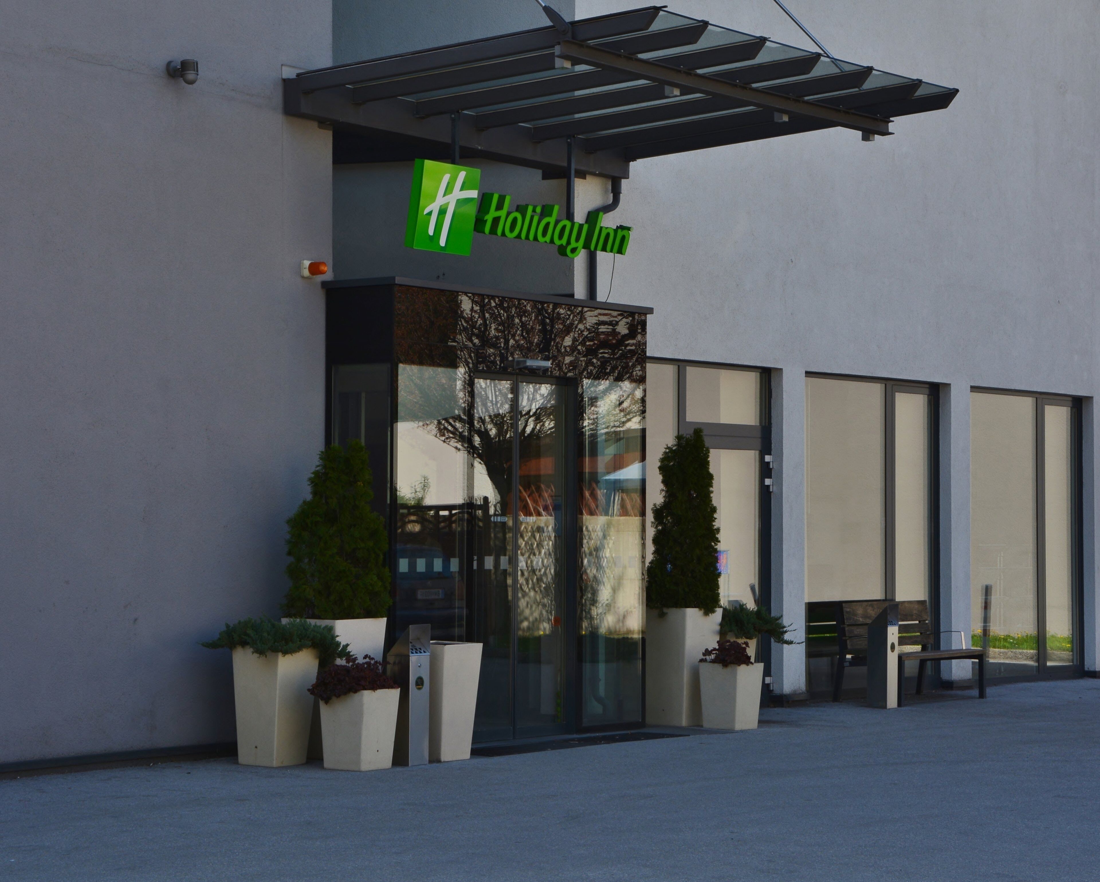 Ingresso di un hotel Holiday Inn con facciata in vetro, posti a sedere all'aperto e piante in vaso.