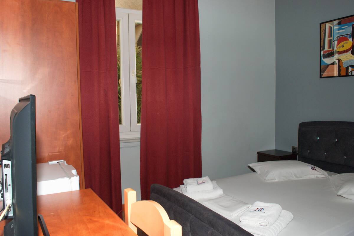 A hotel room with a wooden desk, a bed with white linens, a dark upholstered chair, a nightstand, and a painting on the wall.