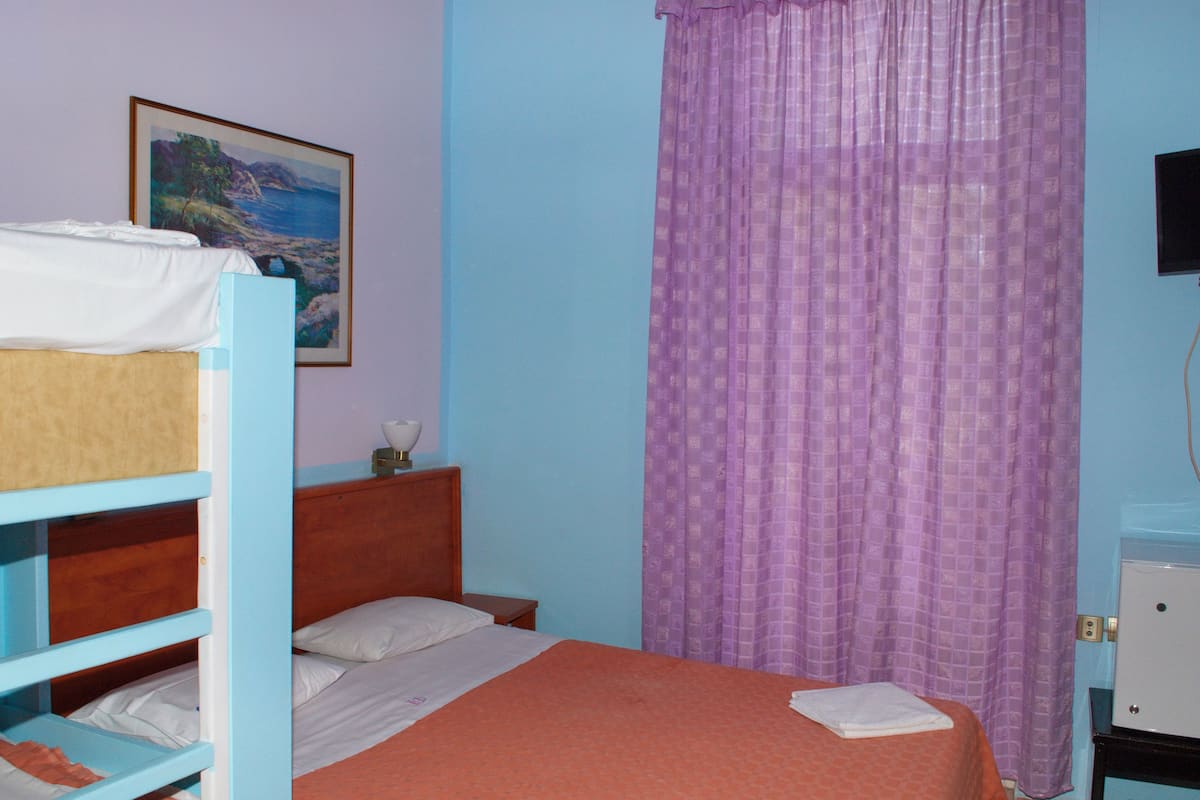 A bedroom with bunk beds, a single bed, a nightstand, a lamp, a painting, a purple curtain, and a television.