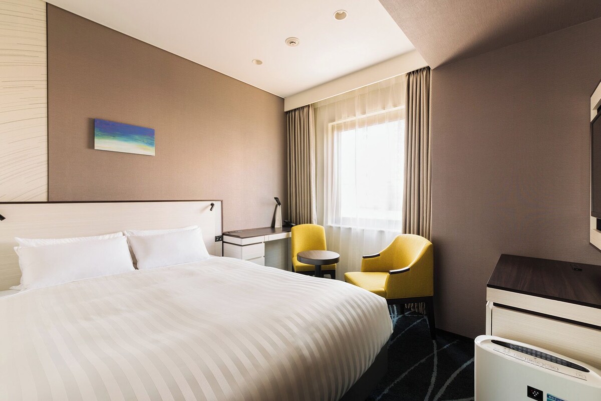 Ein Hotelzimmer mit einem Bett, einem Schreibtisch, einem Stuhl, einem Fernseher und einem Fenster mit Vorhängen.
