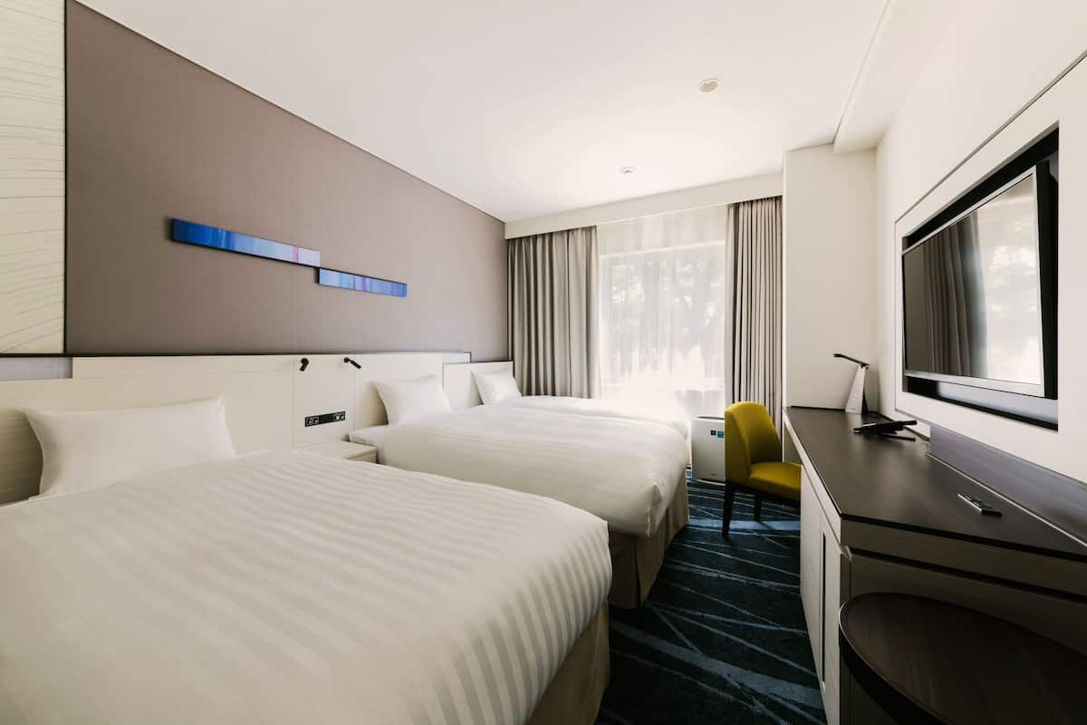 Une chambre d’hôtel avec deux lits, un bureau, une chaise, une télévision et une fenêtre avec des rideaux.
