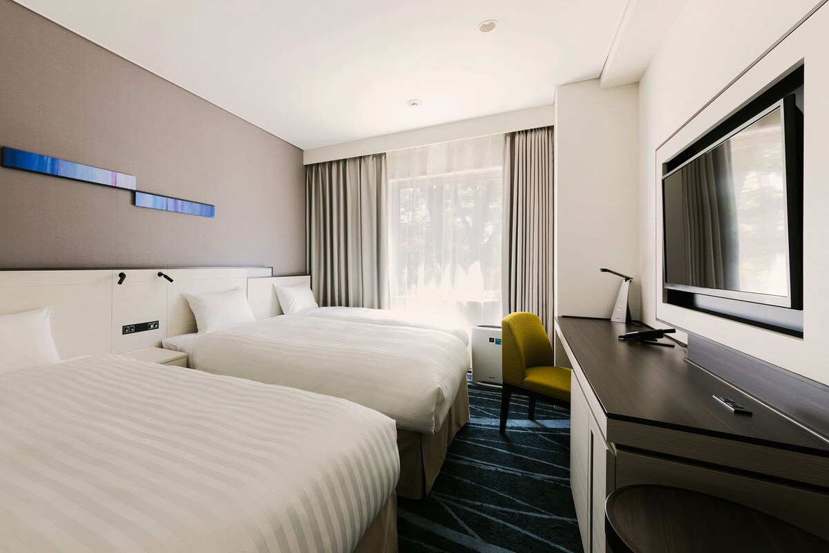 A hotel room with two beds, a desk, a chair, a TV, and a window with curtains.