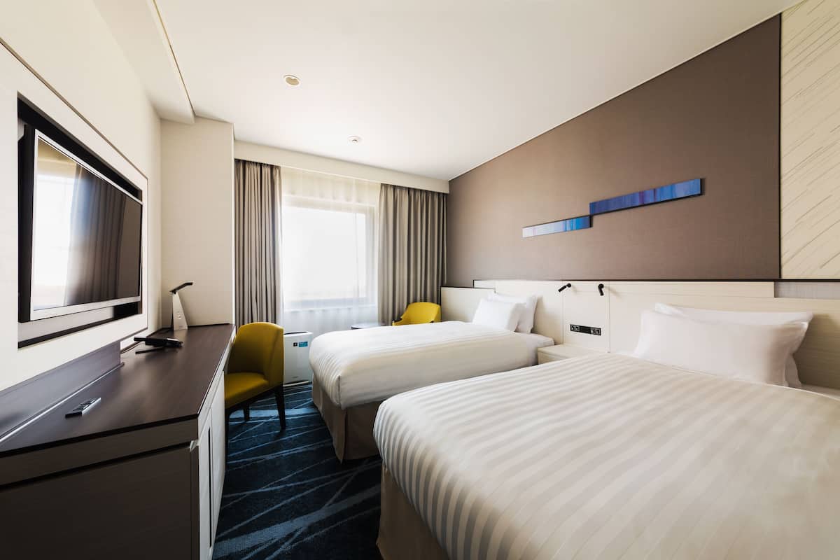 Une chambre d’hôtel avec deux lits, un bureau doté d’un téléviseur à écran plat, une chaise jaune et une grande fenêtre avec des rideaux.