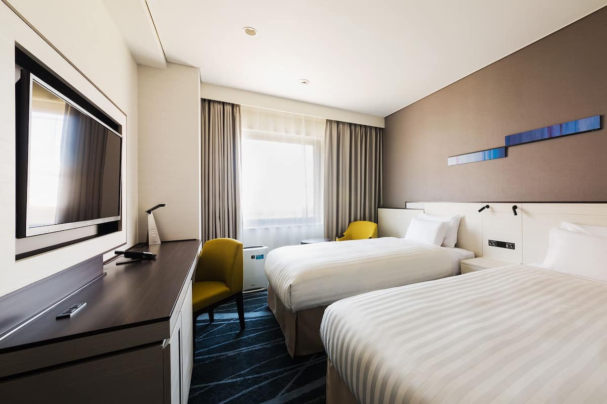 Une chambre d’hôtel avec deux lits, une télévision, un bureau, une chaise et une fenêtre avec des rideaux.