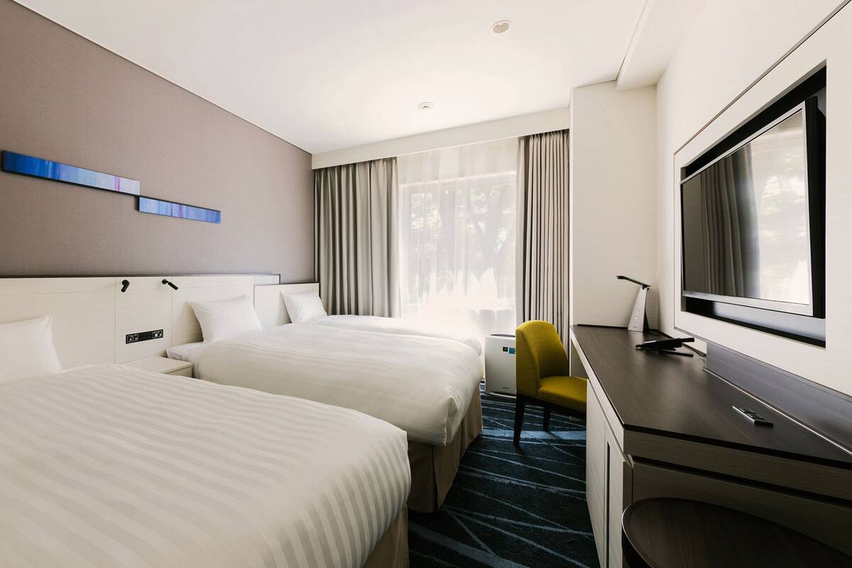 Une chambre d’hôtel avec deux lits, un bureau, une chaise, une télévision et une fenêtre avec des rideaux.