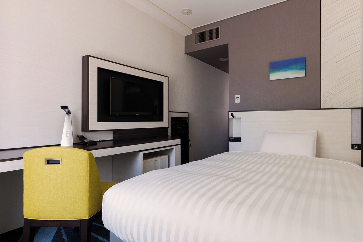 Ein Hotelzimmer mit einem Bett, einem Schreibtisch mit Flachbildfernseher, einem Sessel und einem Bild an der Wand.