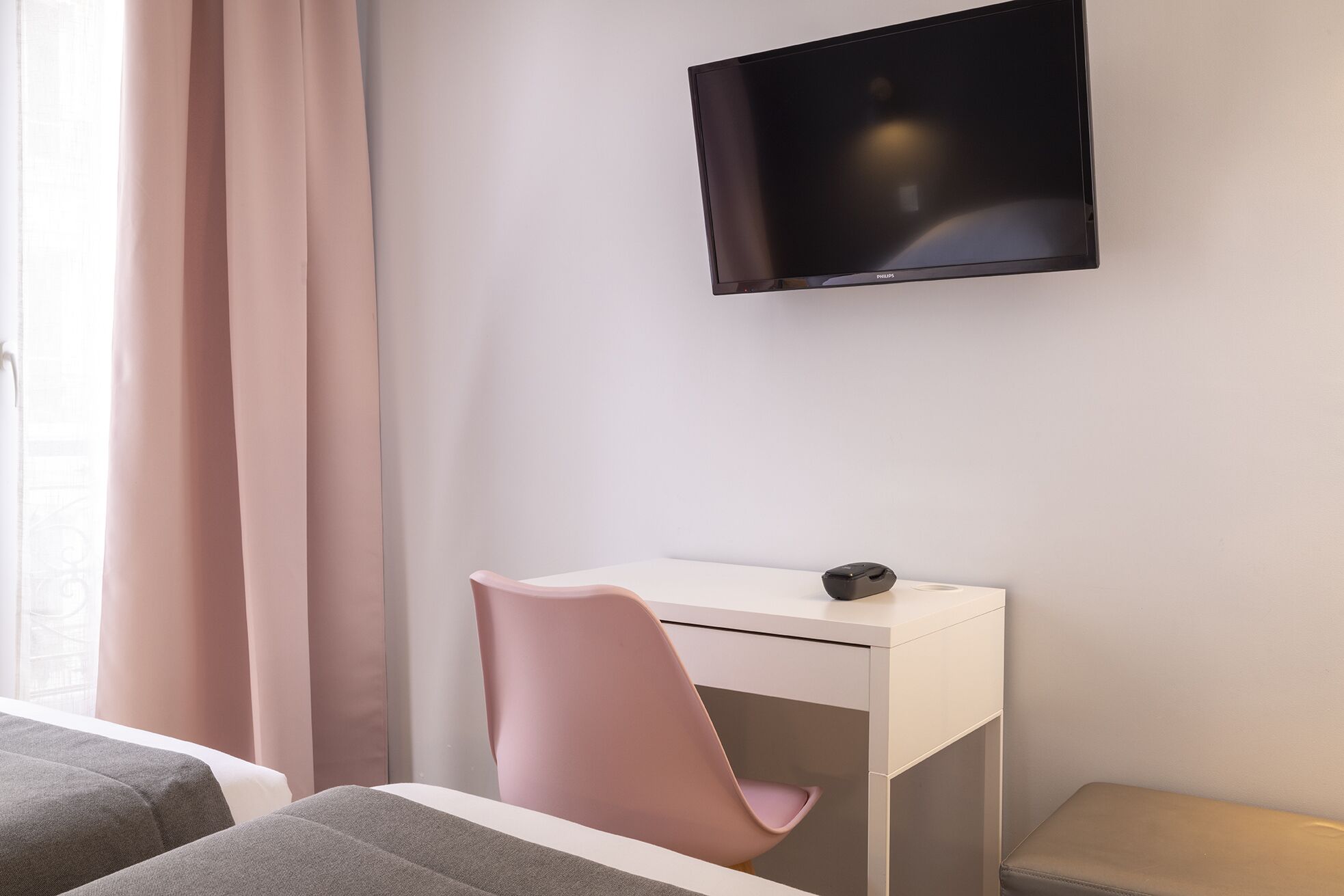 A modern hotel room with a white desk, a pink chair, a flat-screen TV mounted on the wall, and a window with curtains.