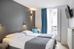 A hotel room with two beds, a desk, a TV, and a window with a view of a building.