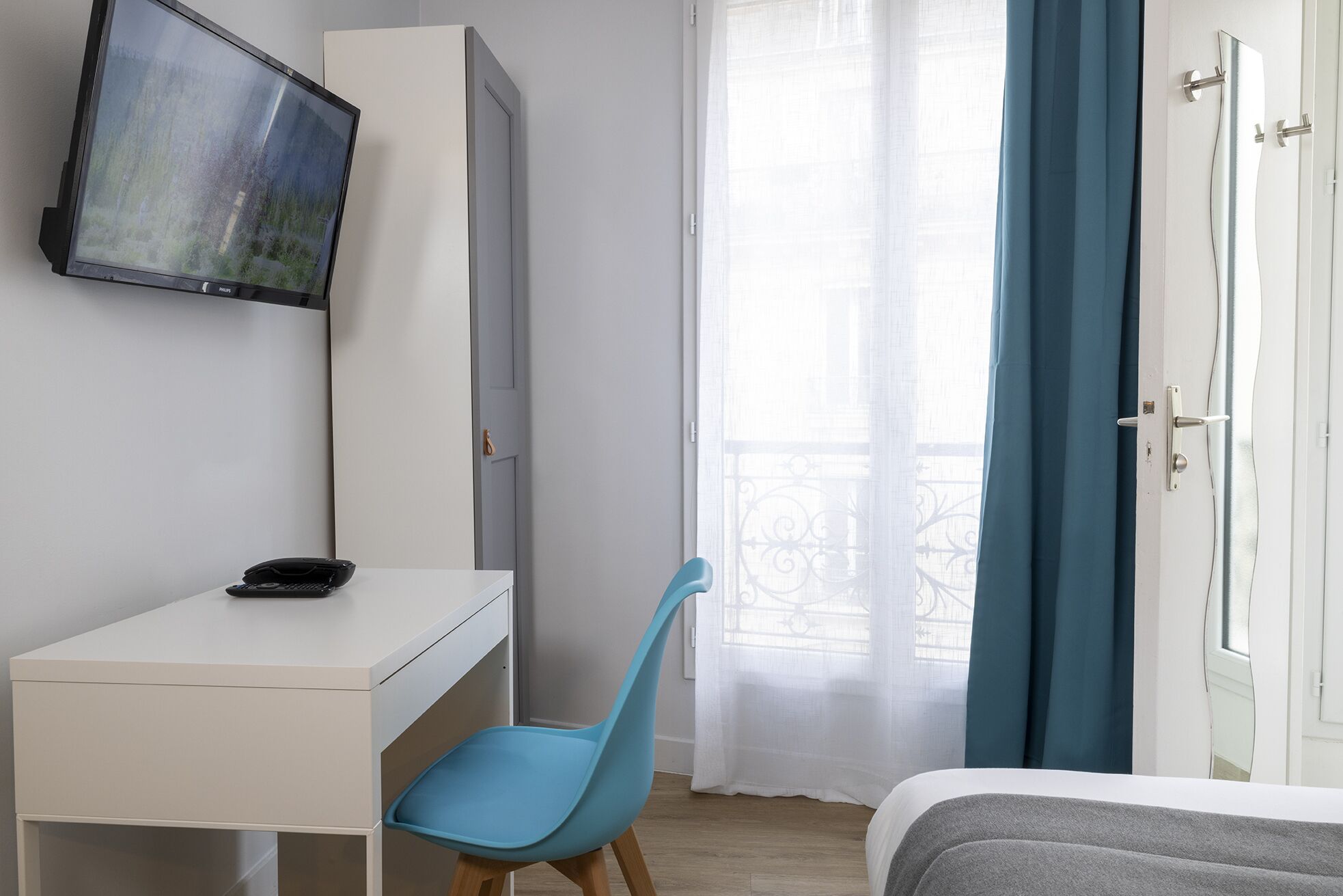Une chambre à coucher avec un lit, un bureau avec une chaise, un téléviseur fixé au mur et une fenêtre avec des rideaux.