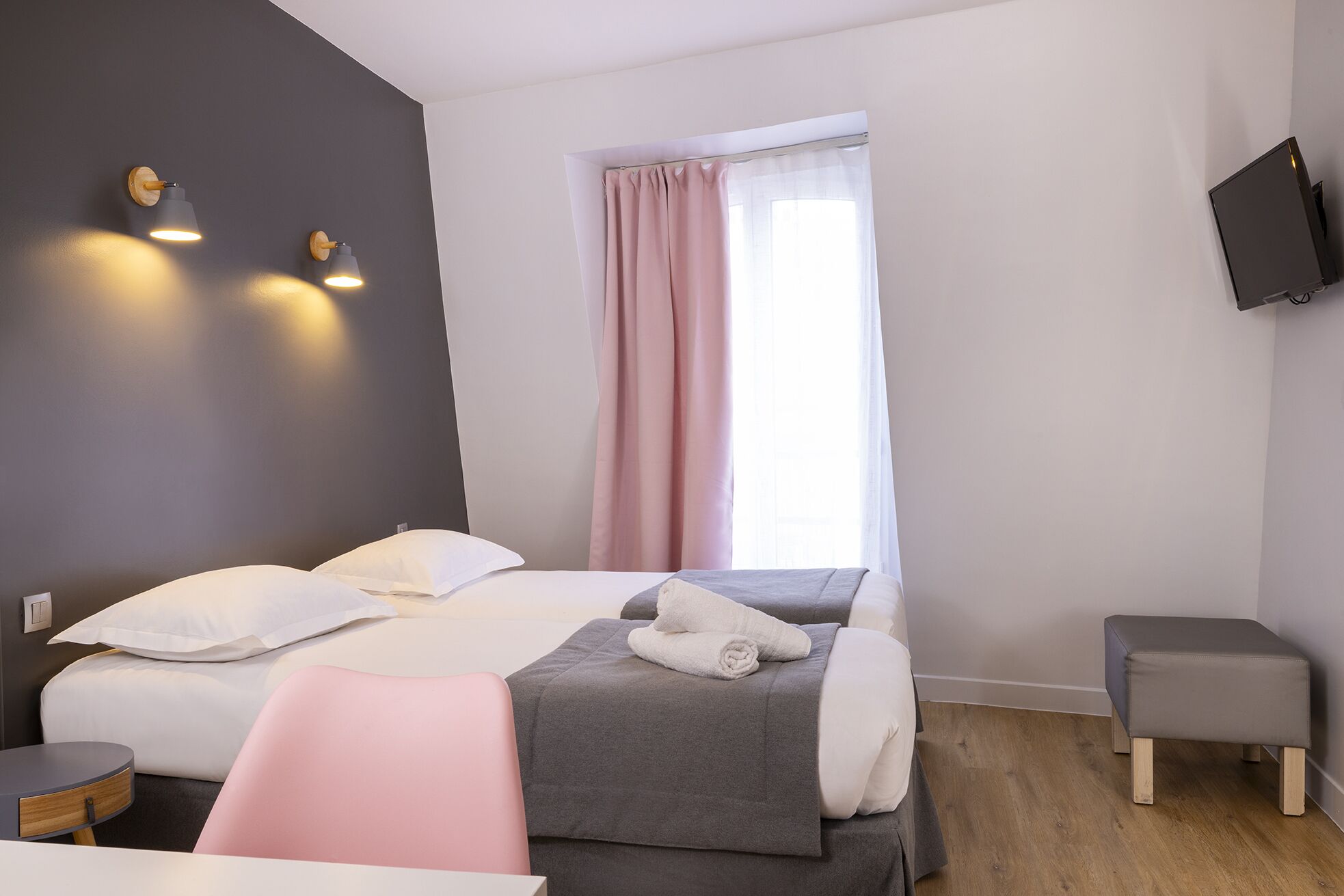 A modern hotel room with a bed, bedside tables, a chair, a TV, and a window with curtains.