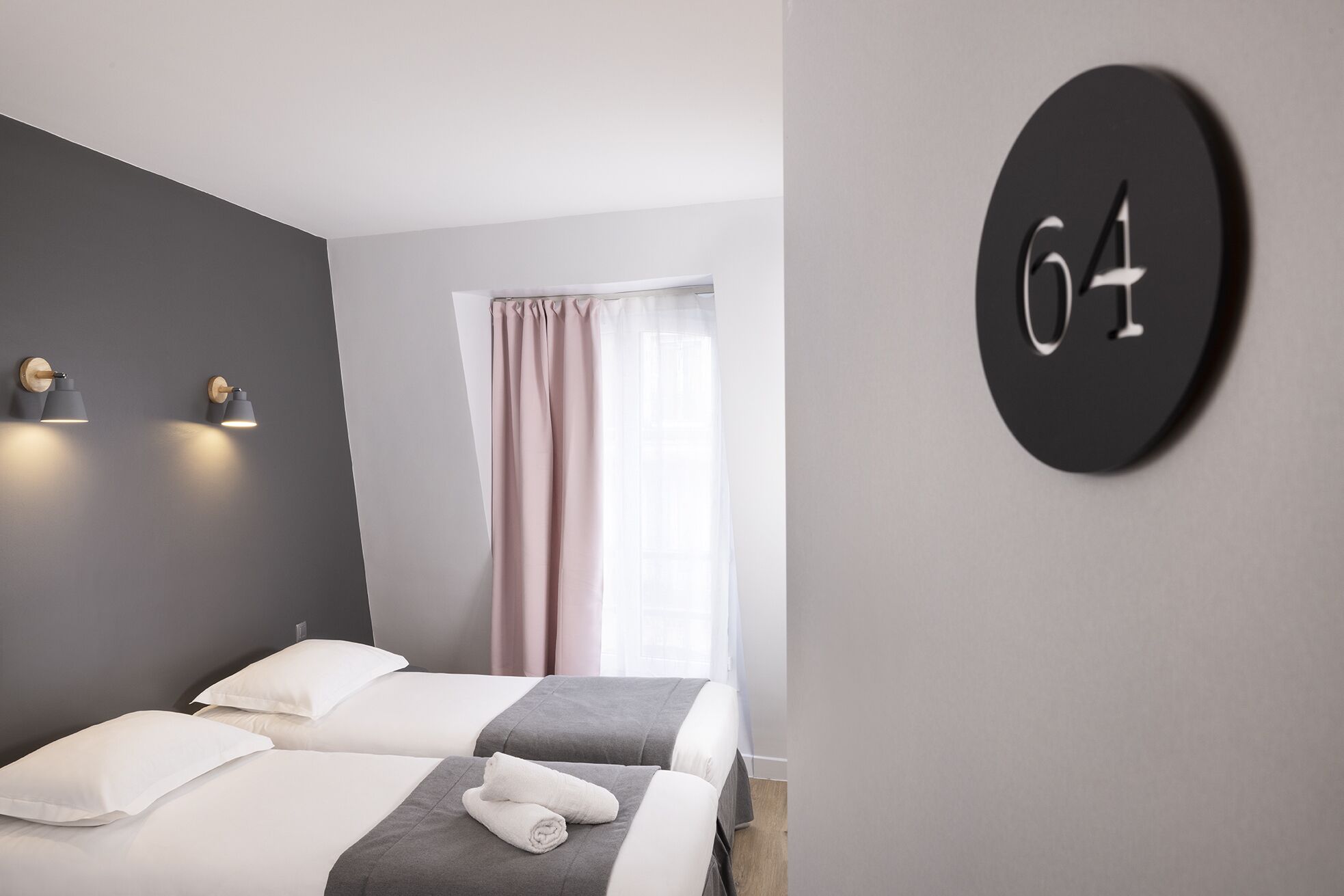 A hotel room with two beds, a large window with sheer curtains, and a wall-mounted number sign.