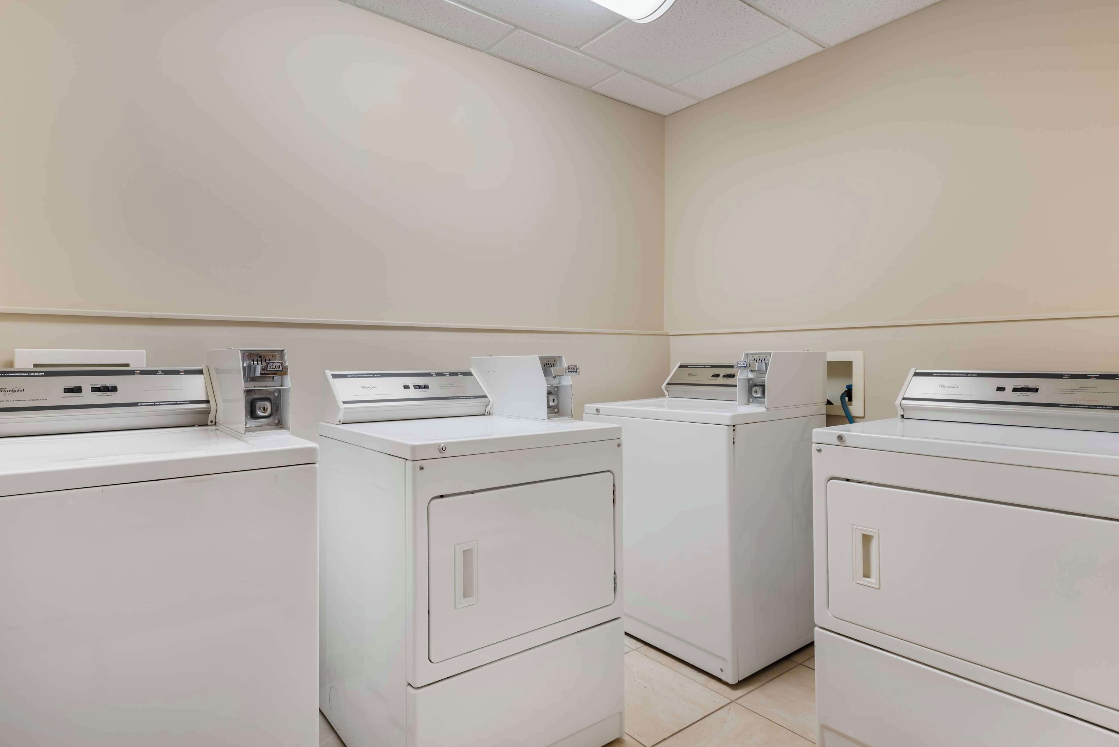 A laundry room with four white washing machines and dryers, a control panel, and a paper towel dispenser.