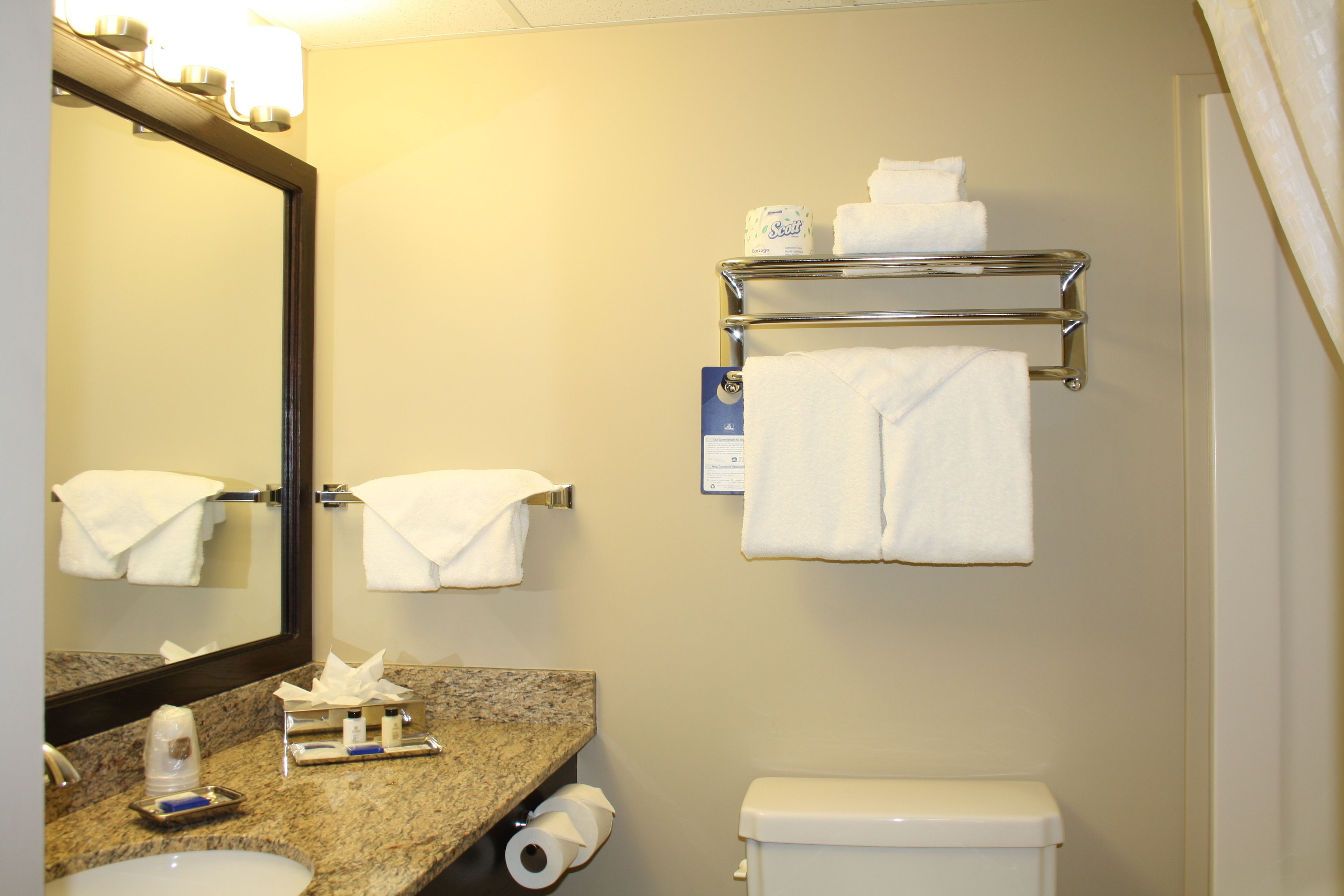A bathroom with a granite countertop, a sink, a toilet, a towel rack with towels, a mirror, and a paper towel dispenser.
