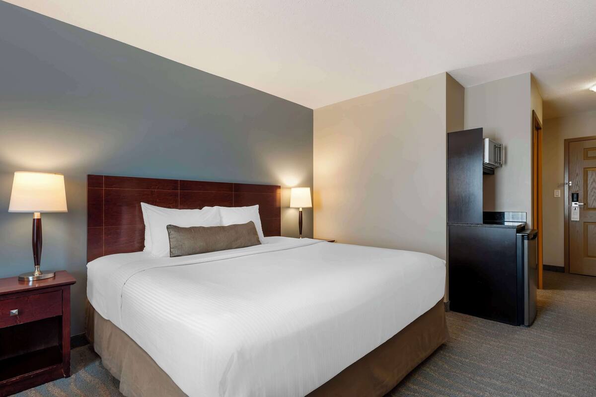 Une chambre d’hôtel avec un grand lit, deux lampes de chevet, une tête de lit en bois foncé, un petit réfrigérateur et une porte avec une serrure.
