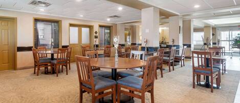 A spacious dining area with multiple round tables and wooden chairs, featuring a carpeted floor and recessed ceiling lighting.